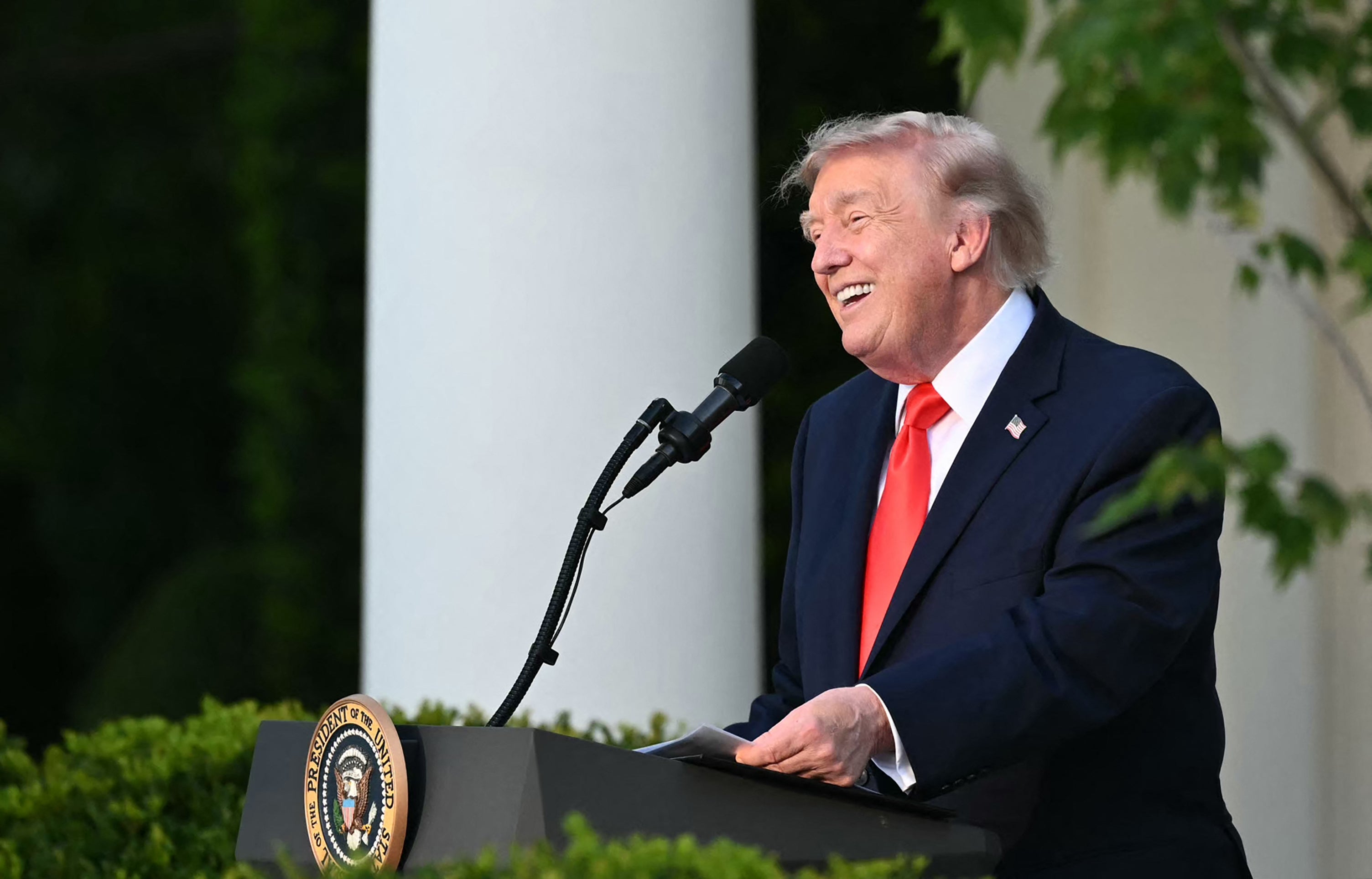 A antheral successful a suit and necktie speaks astatine a podium with the statesmanlike seal, outdoors, smiling arsenic helium addresses an audience