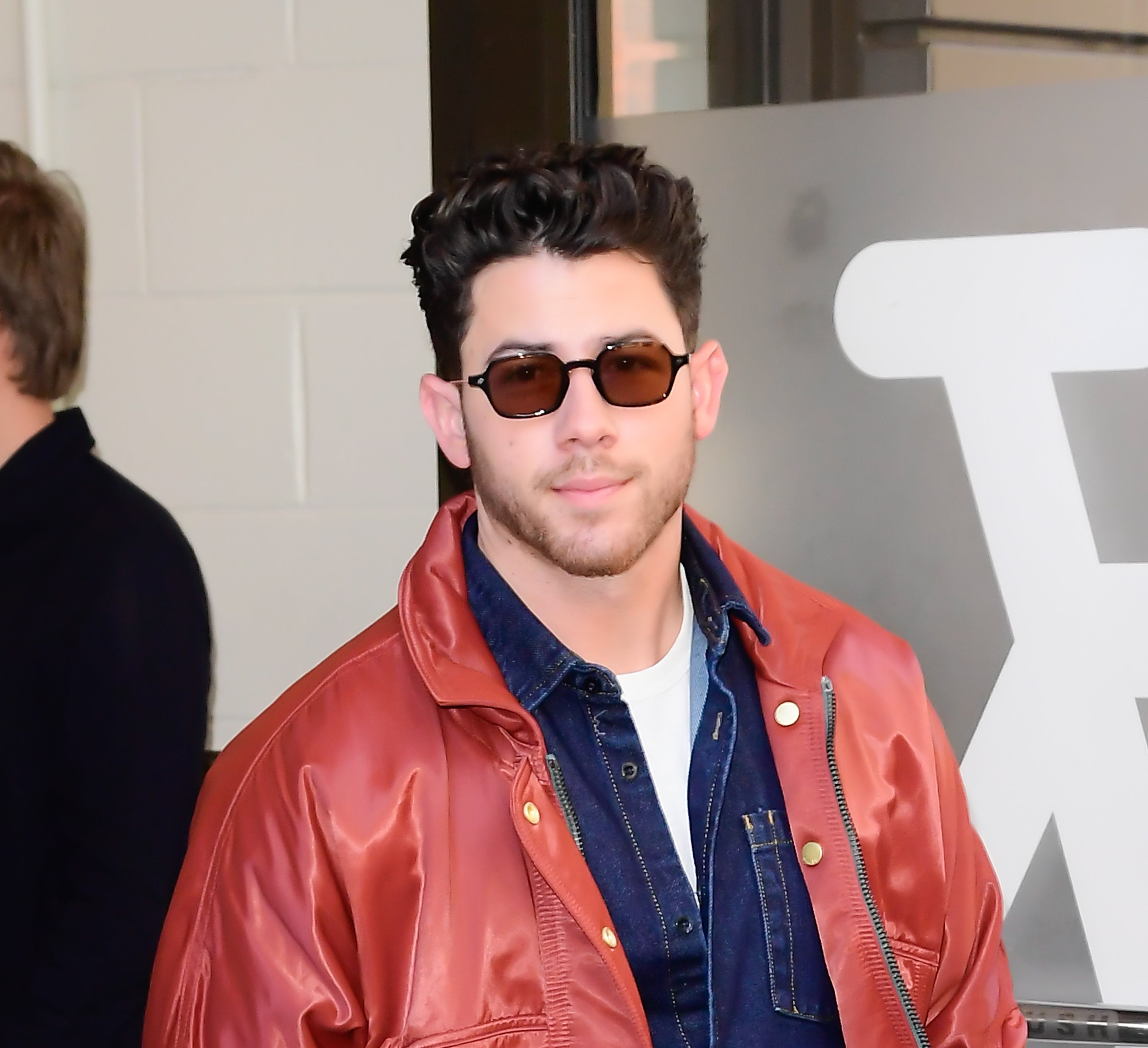 A person is wearing a leather jacket over a denim shirt and sunglasses, standing in front of a building entrance