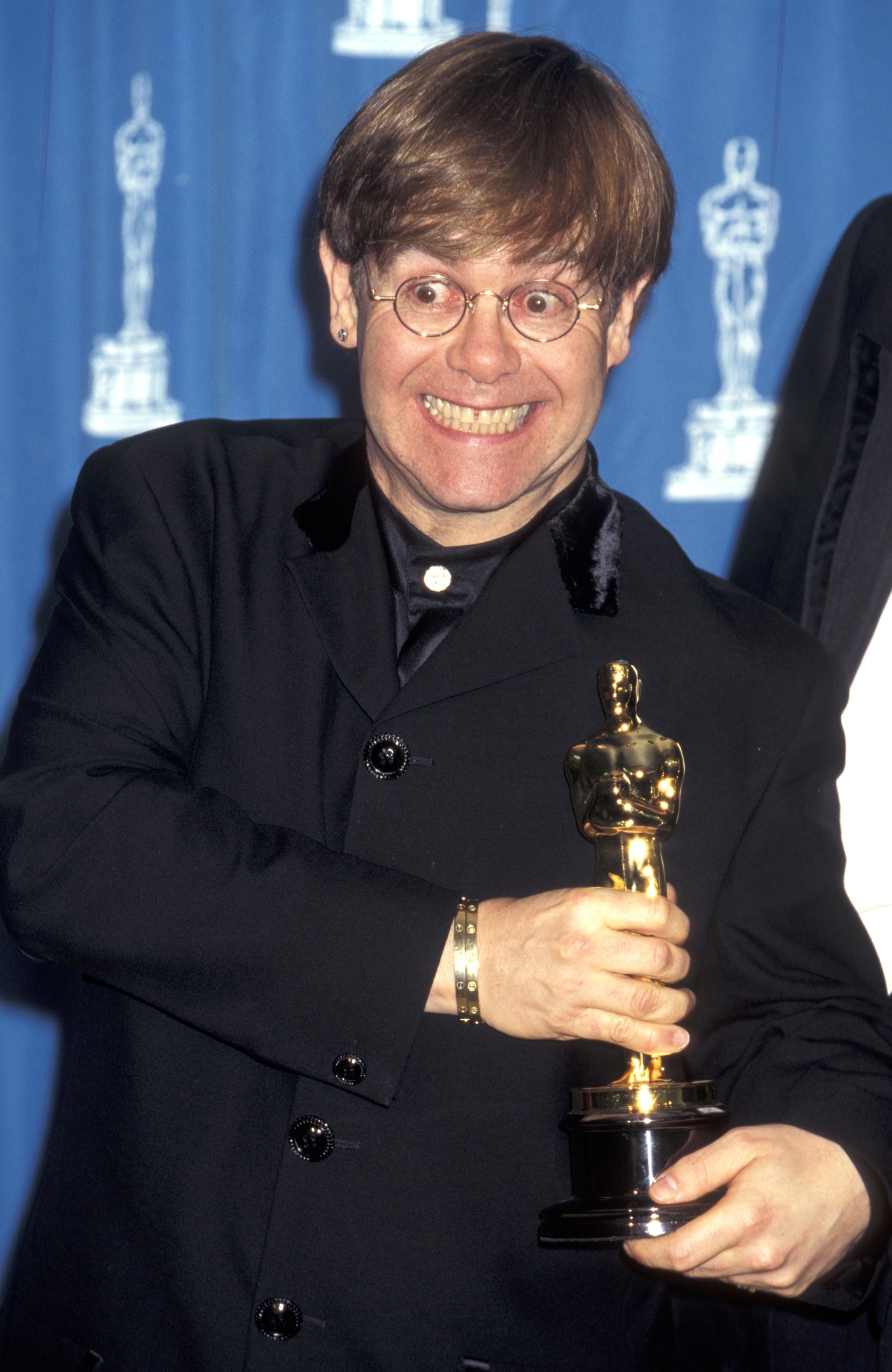 A person with a joyful expression holds an Oscar statuette, dressed in a classic black suit