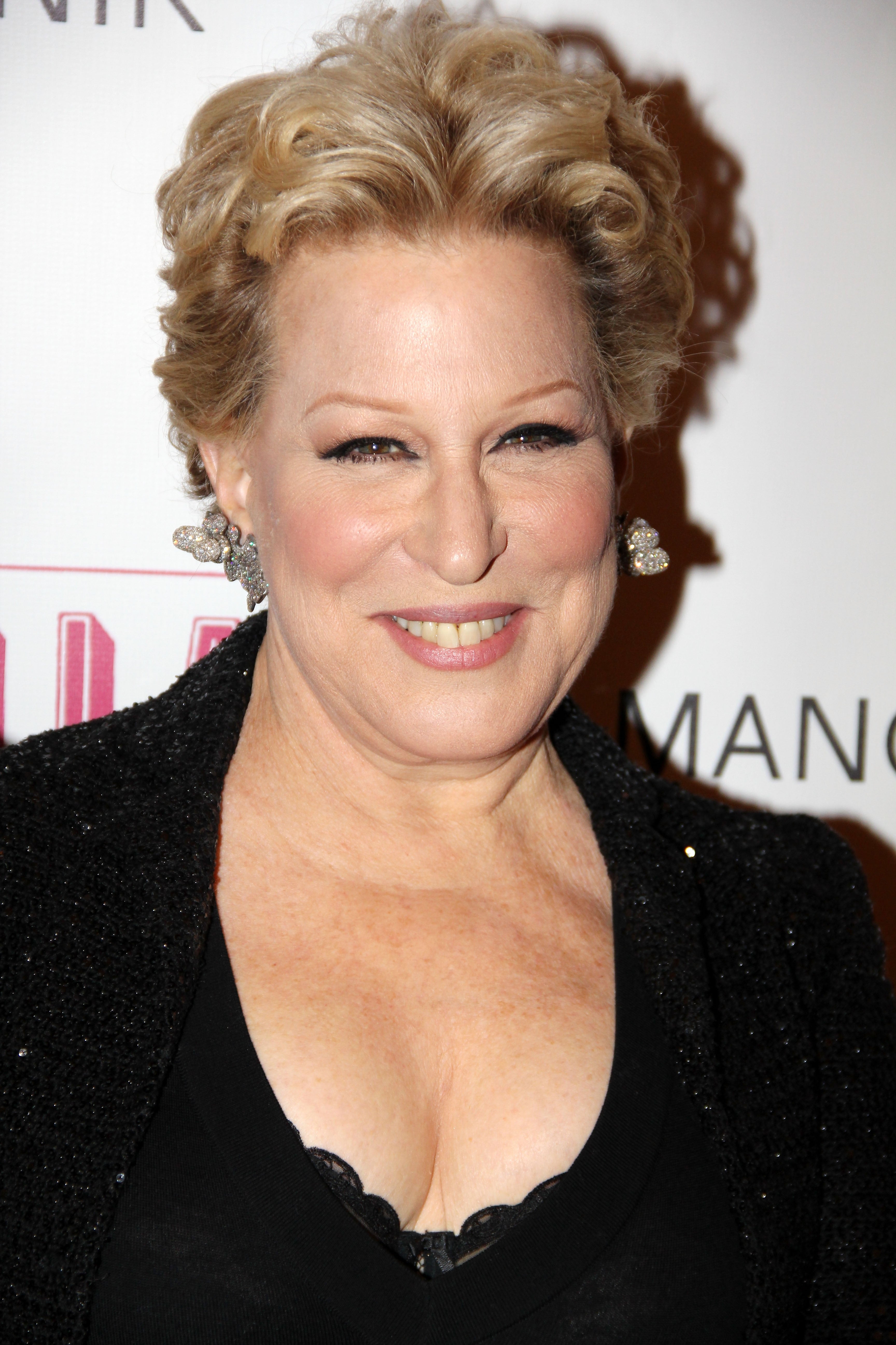 Bette Midler smiles at an event, wearing a black outfit with a deep neckline and sparkling earrings