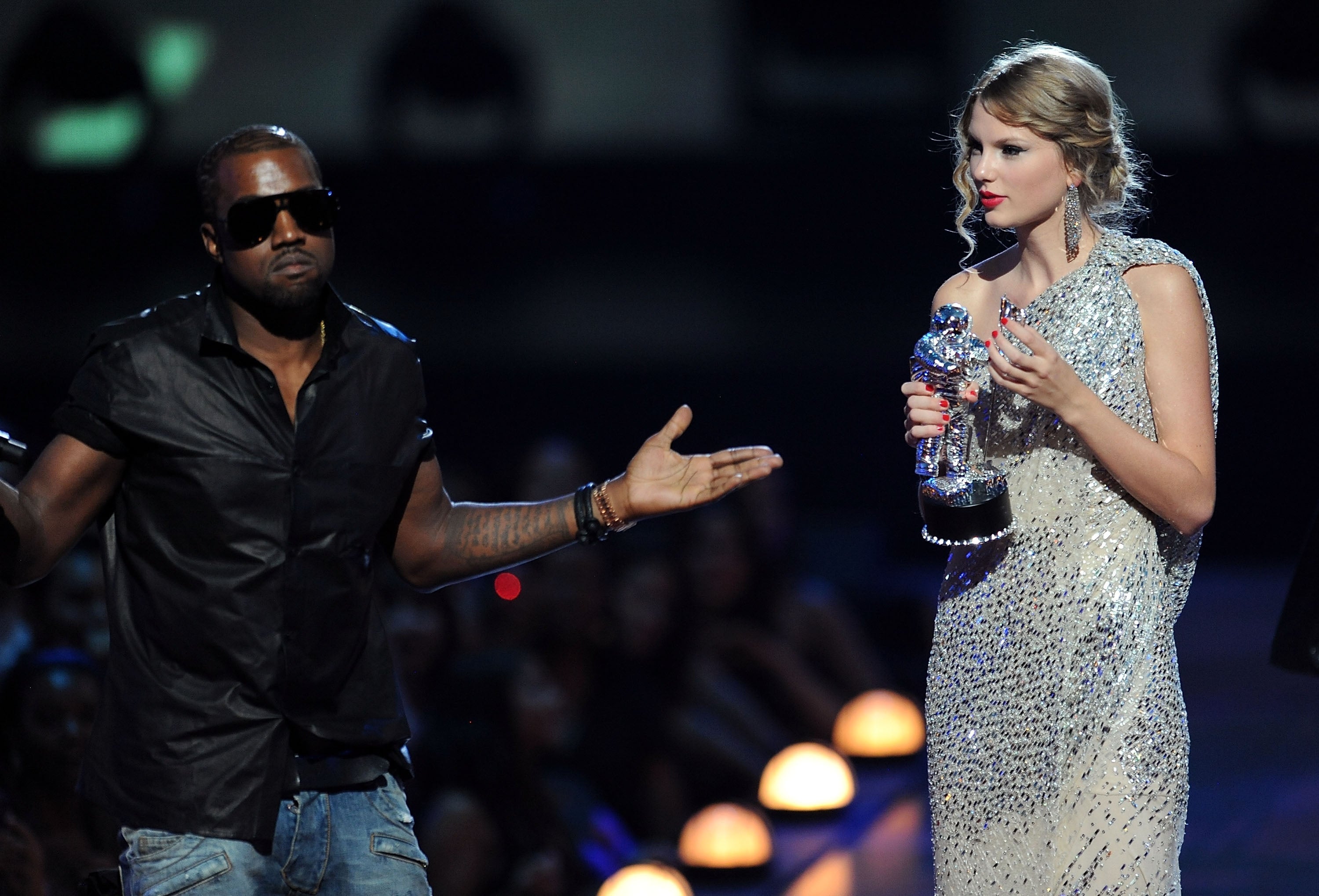 Kanye West interrupts Taylor Swift's award speech on stage. She's in a sequined dress, holding a trophy and looking surprised