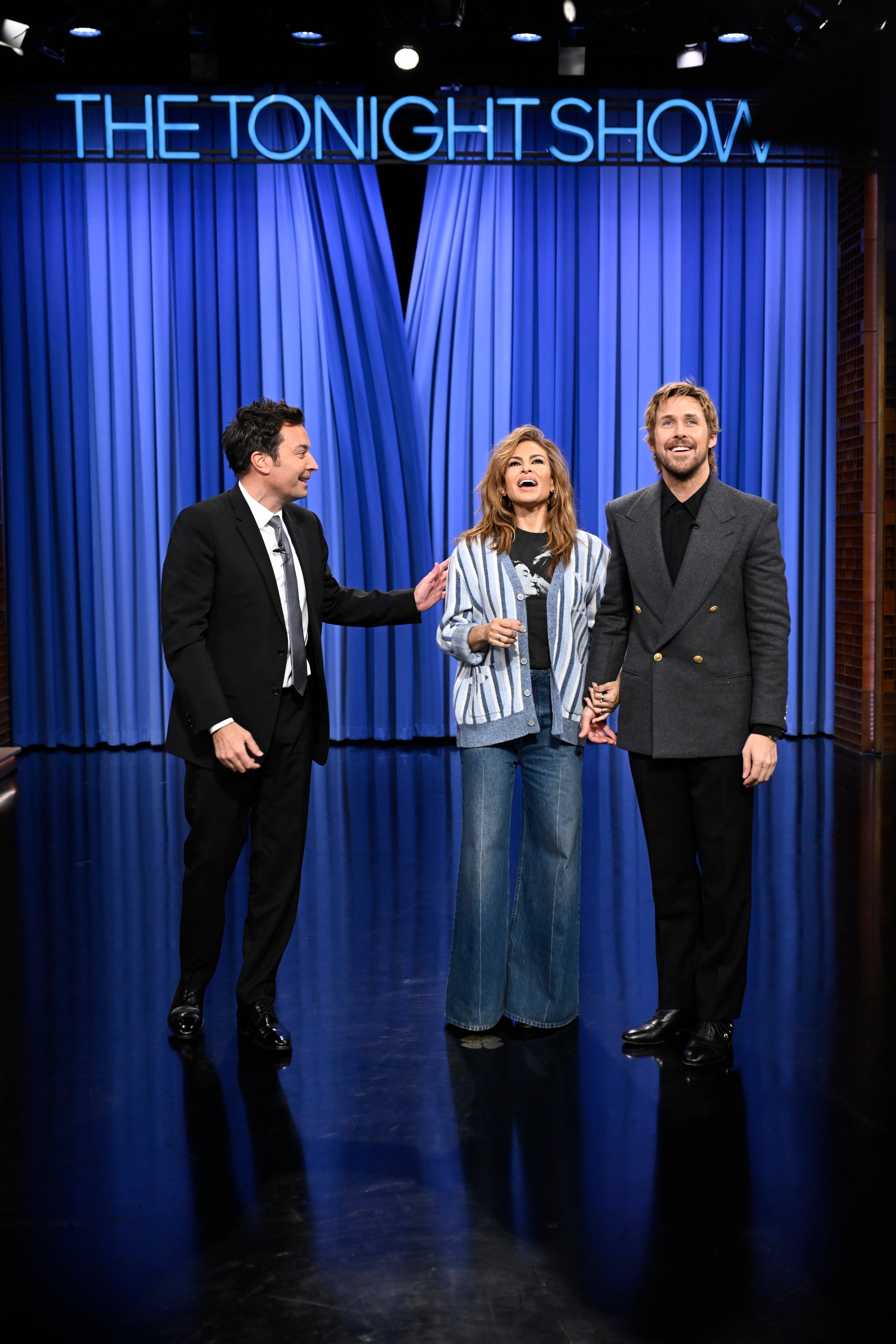 Three people on a talk show stage, one in a suit, another in flared jeans and striped top, and the third in a tailored coat