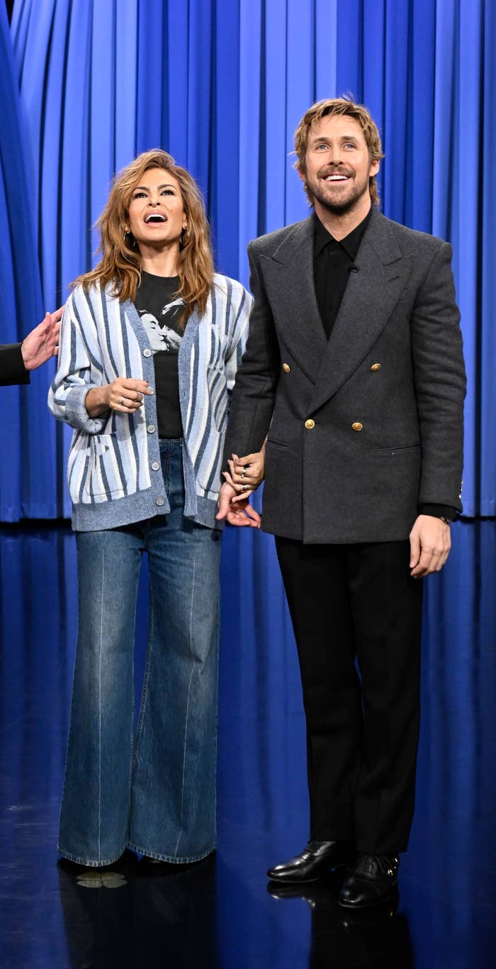Three people on a talk show stage, one in a suit, another in flared jeans and striped top, and the third in a tailored coat