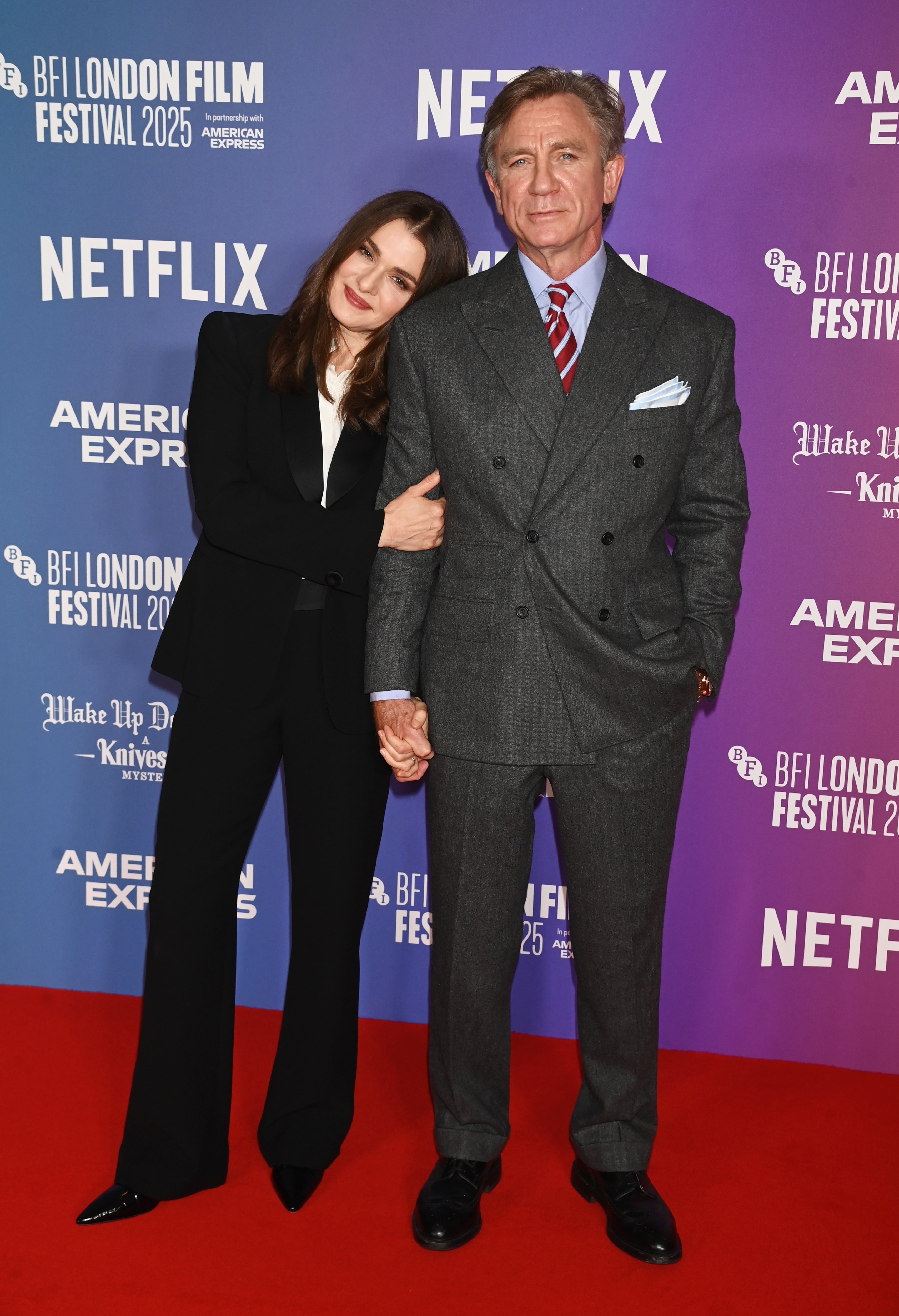 Two people pose on the red carpet at a film festival. The woman wears a black suit, and the man wears a grey suit with a red tie