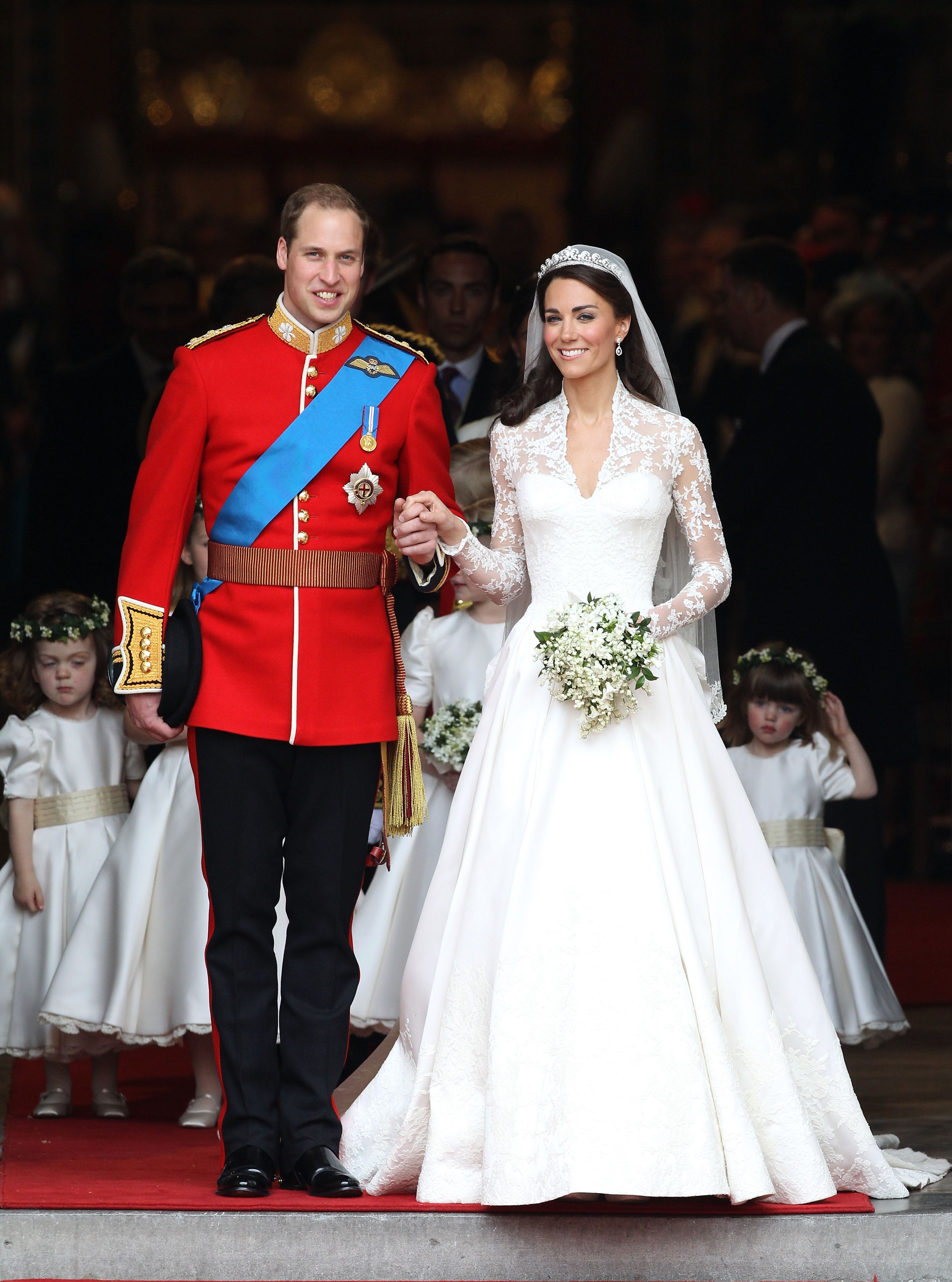 A couple in formal attire, with the man in a military uniform and the woman in a lace wedding dress, stands on a red carpet, surrounded by bridesmaids