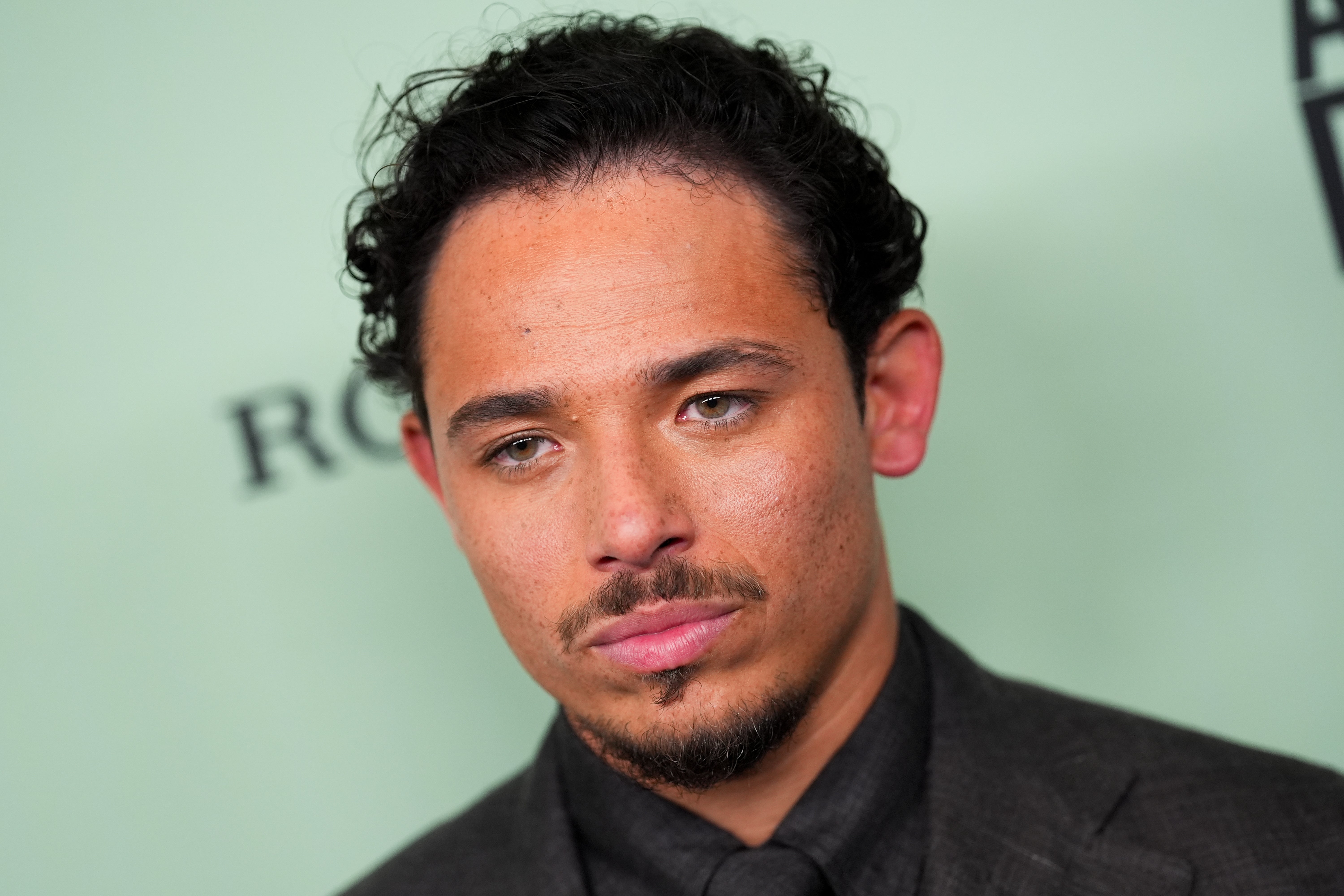 Anthony Ramos scowls as he attends an event with a blurred mint green background