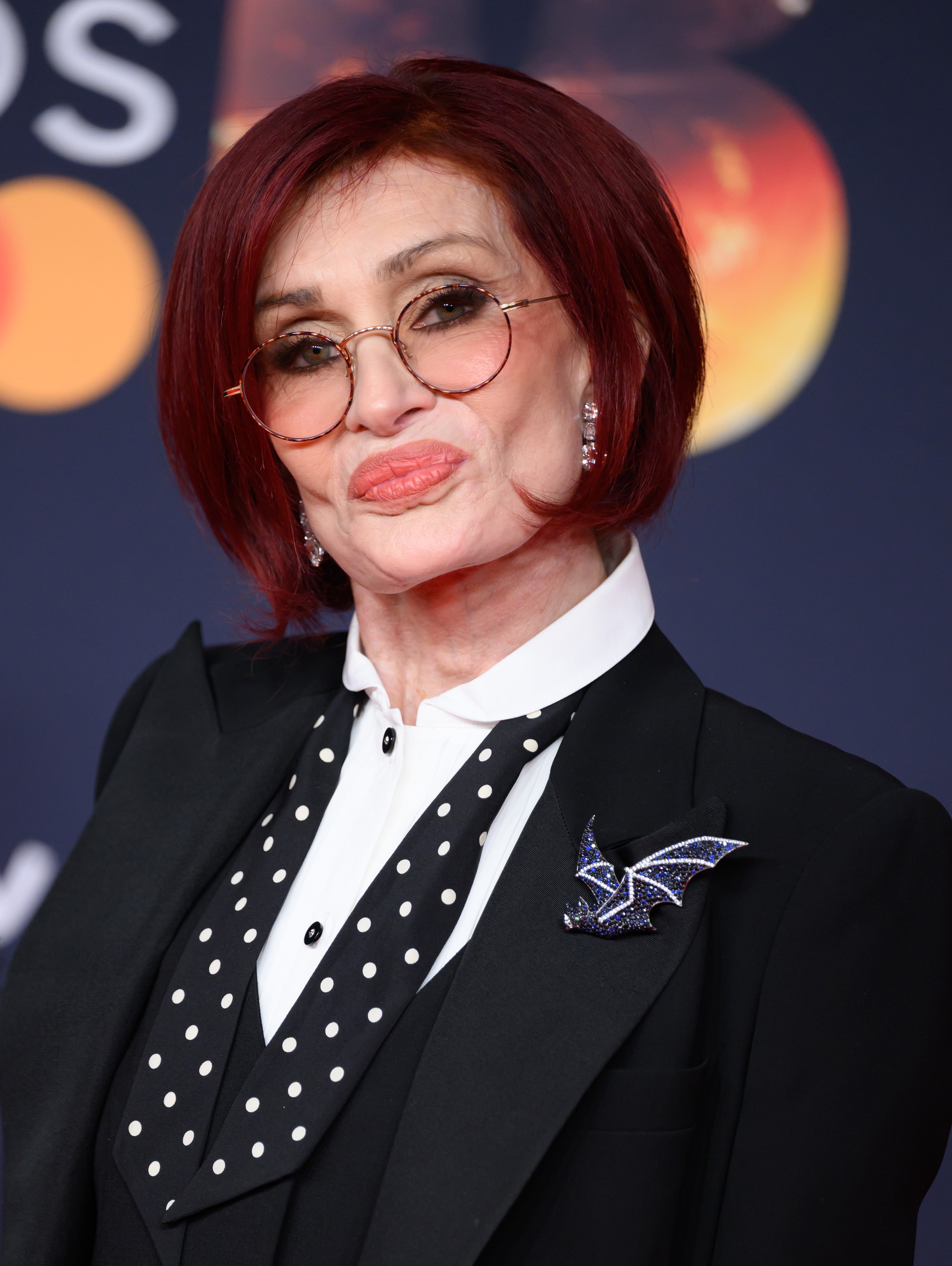 Sharon Osbourne in a polka-dot shirt, a black blazer with a brooch, and round glasses, poses on a red carpet