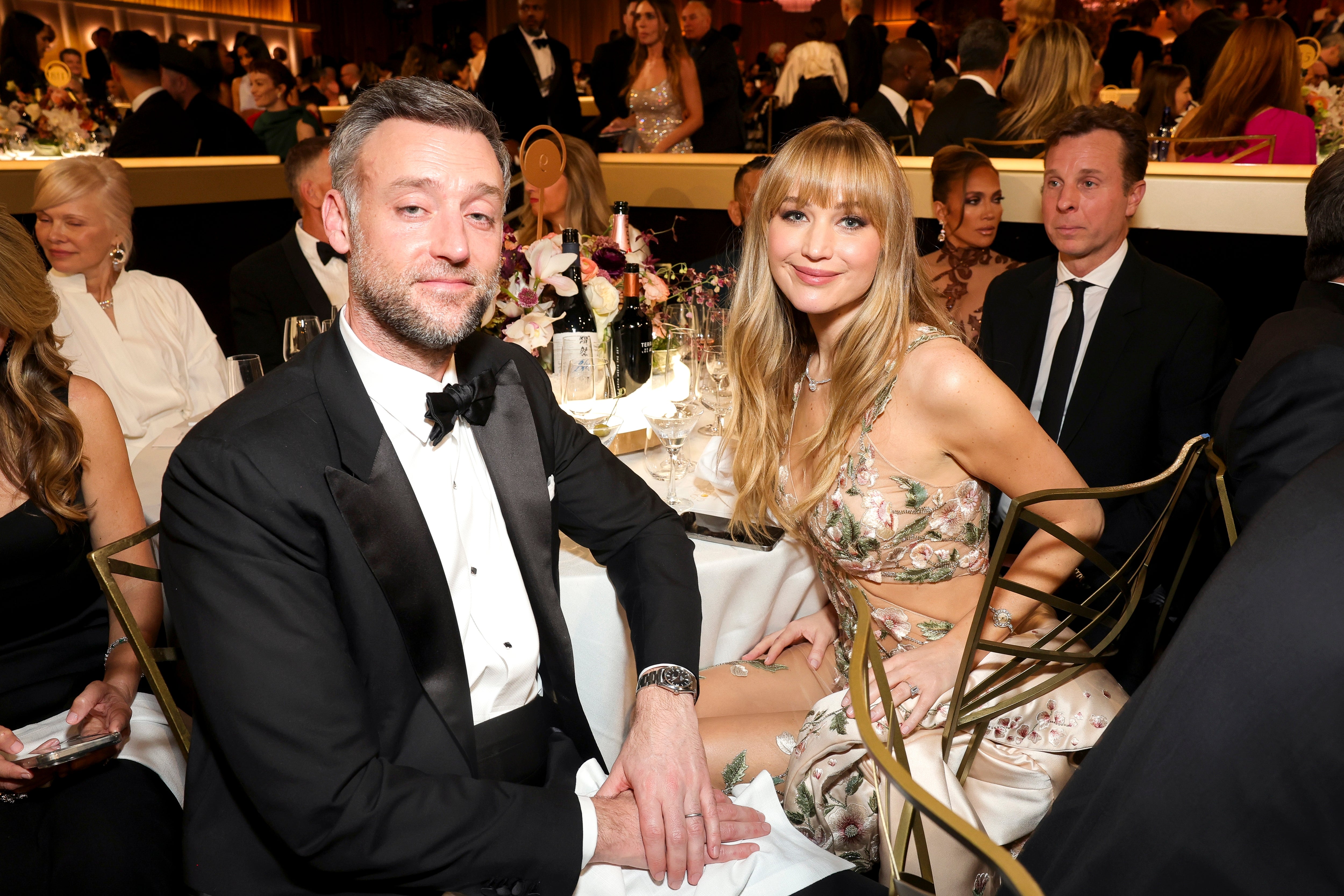 Two people sit at an elegant event table, with others in formal attire in the background. One wears a tuxedo, the other a stylish floral gown