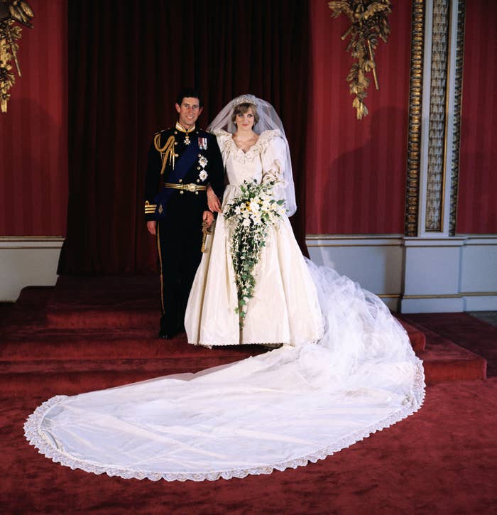 A bride in an elaborate wedding gown and veil stands beside a groom in a formal military uniform, posing indoors on a red carpeted stairway