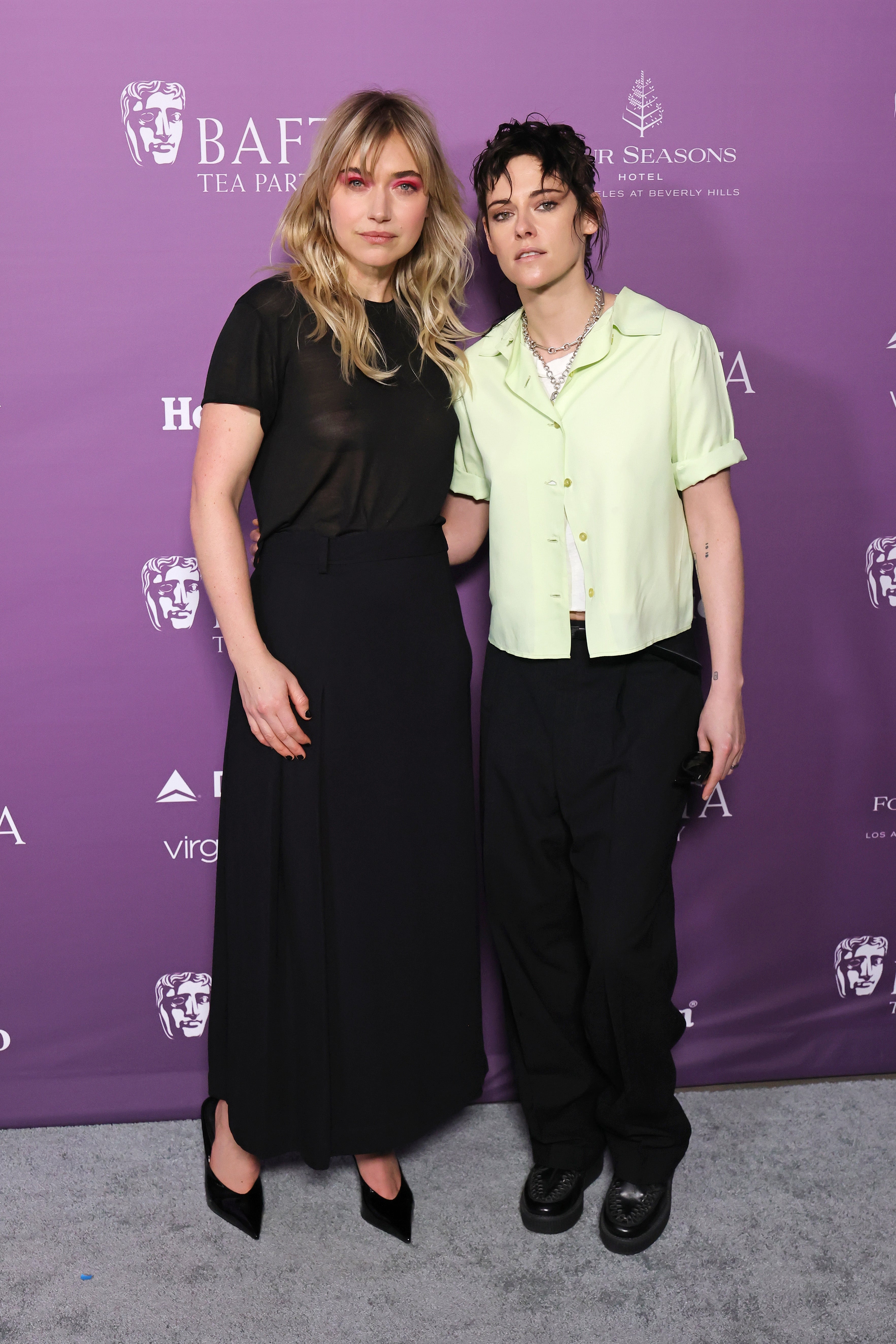 Two individuals stand together at a BAFTA event. One wears a black top and skirt, the other a light shirt and dark pants, posing for photos