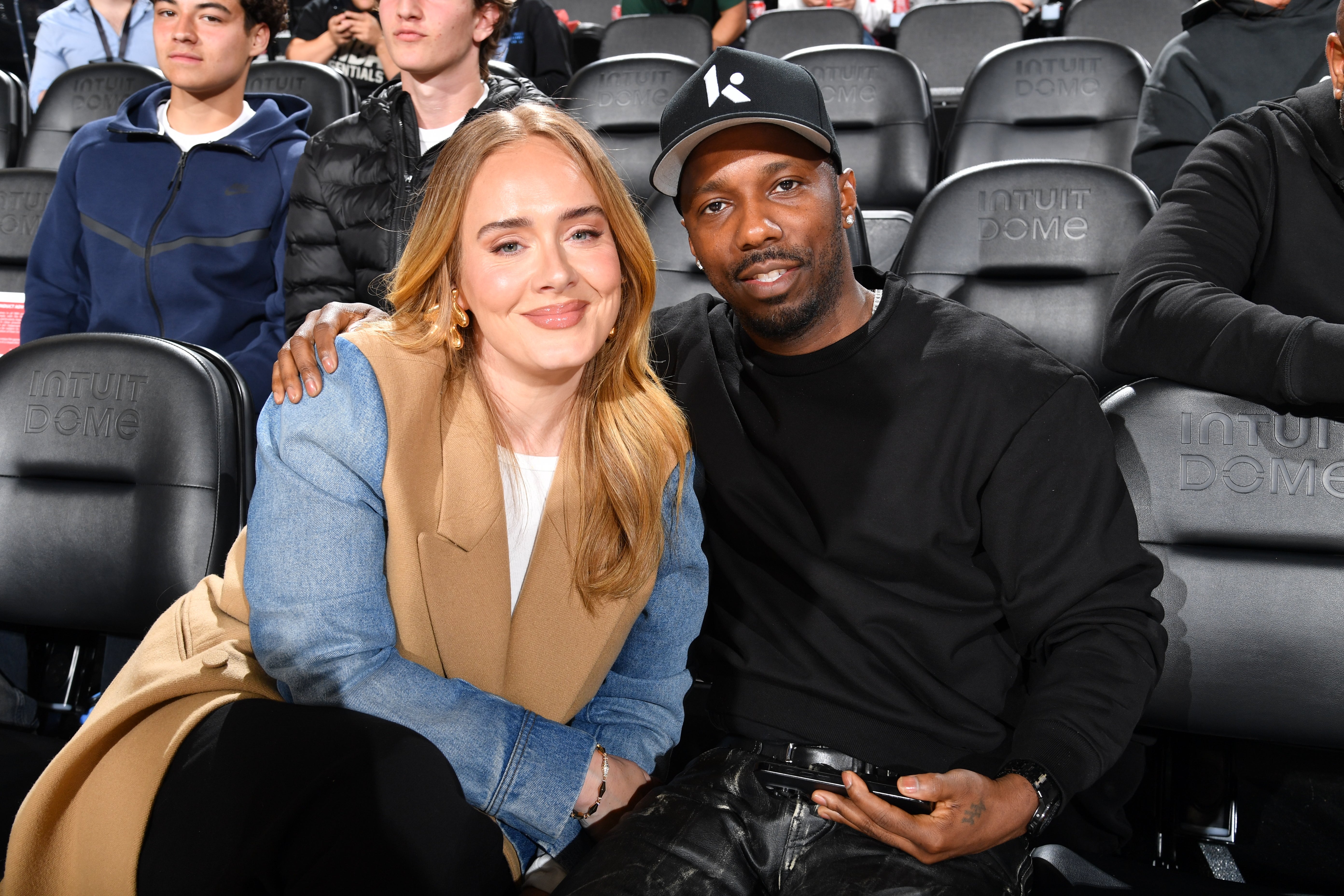 Two people sitting courtside at a sporting event, one wearing a stylish long coat and the other wearing casual attire