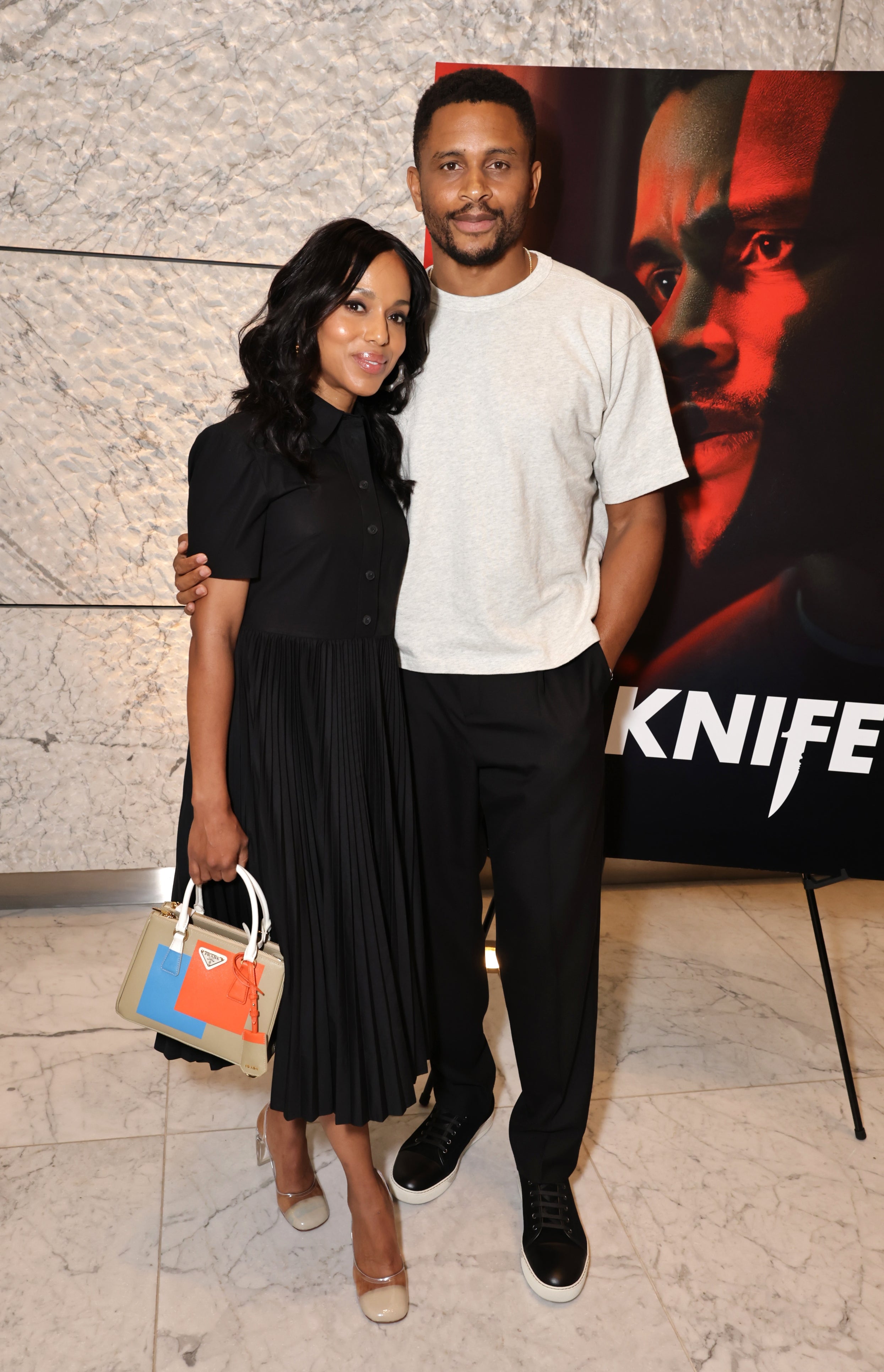 A woman in a pleated dress and a man in casual attire pose together at a media event, with a promotional poster in the background