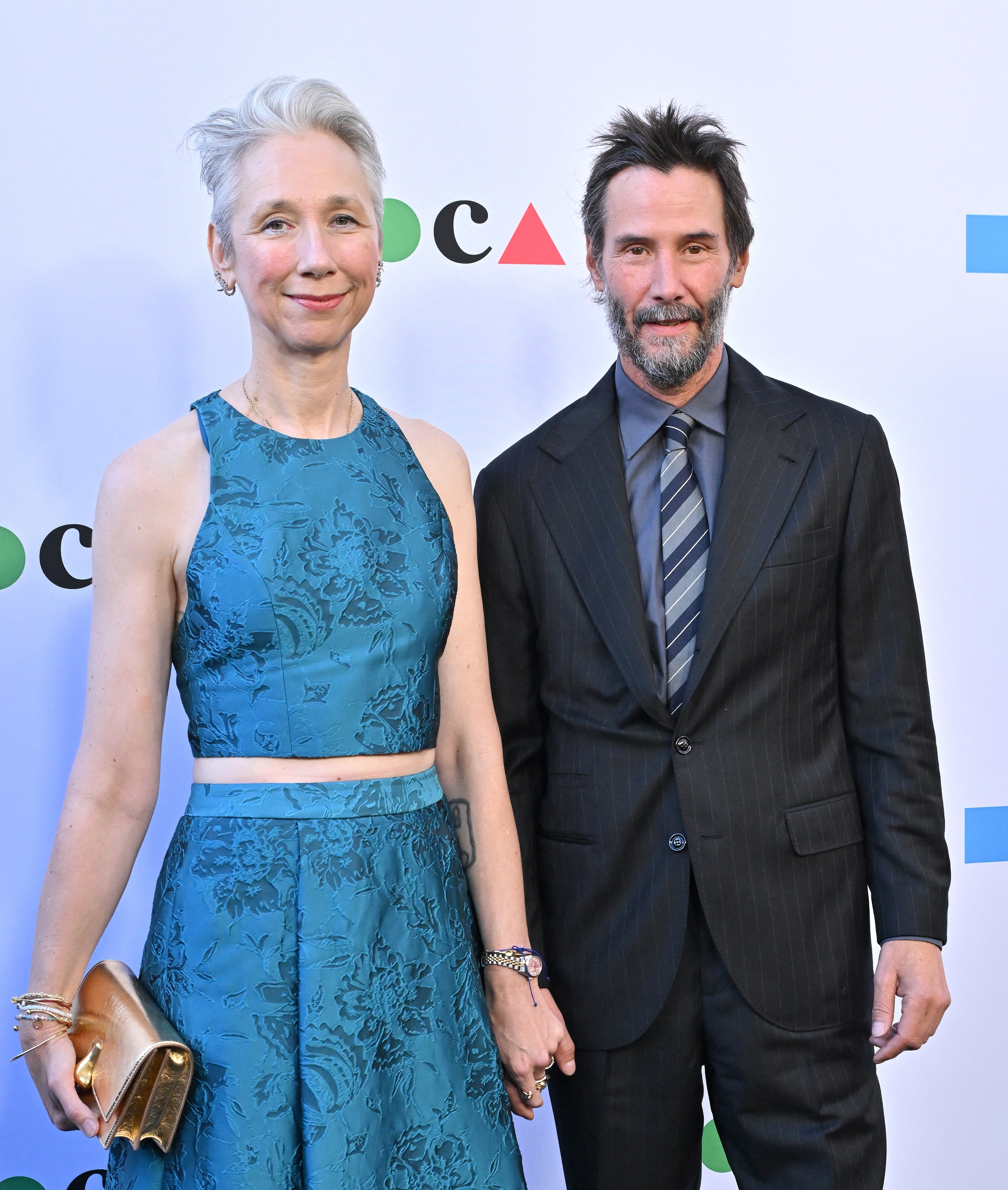 Two individuals on a red carpet; one wears a sleeveless floral dress, the other a dark suit with a striped tie, holding hands and smiling
