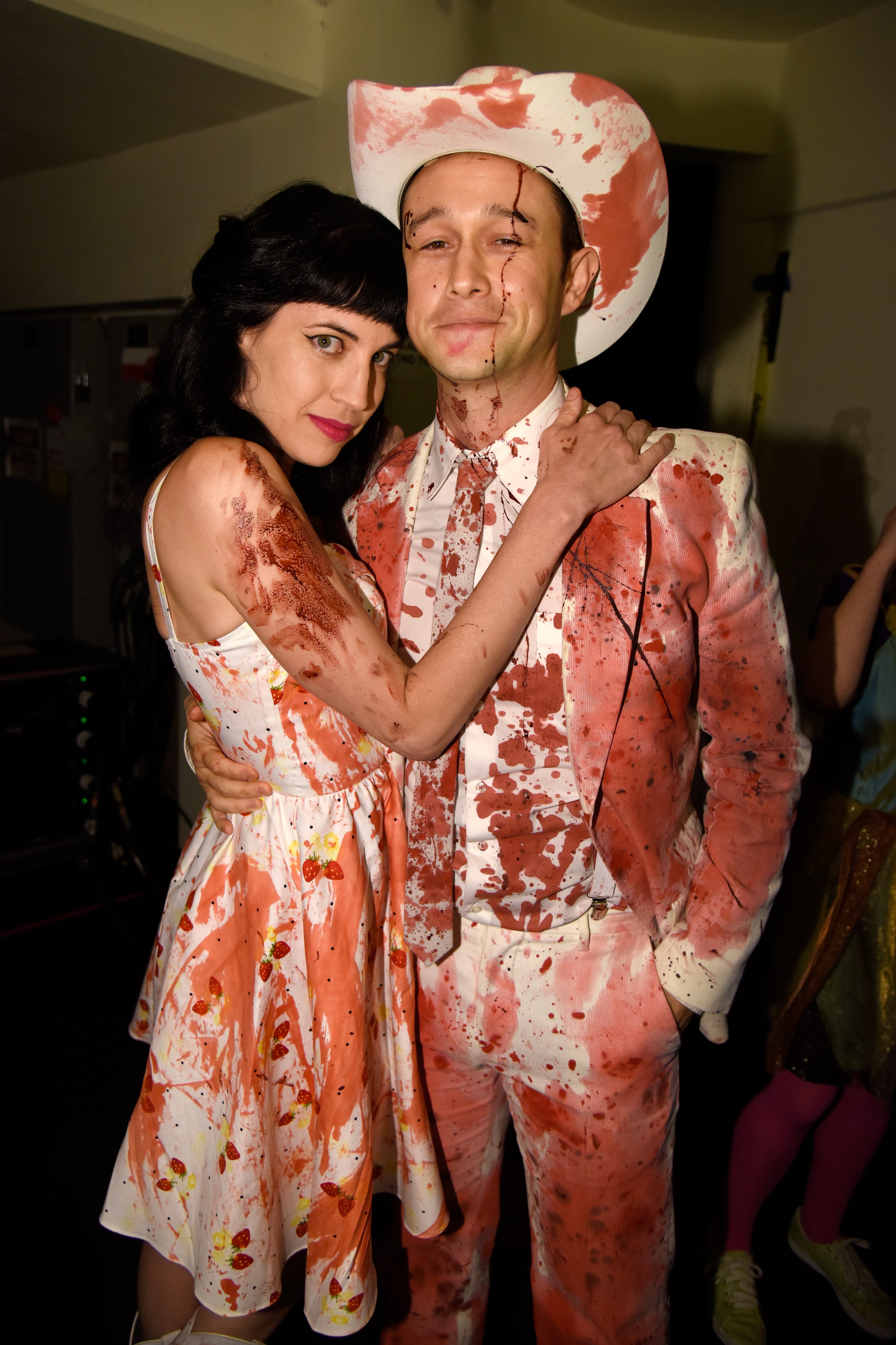 Two people on a red carpet wearing matching splattered outfits; one in a dress and the other in a suit and cowboy hat, creating a themed ensemble