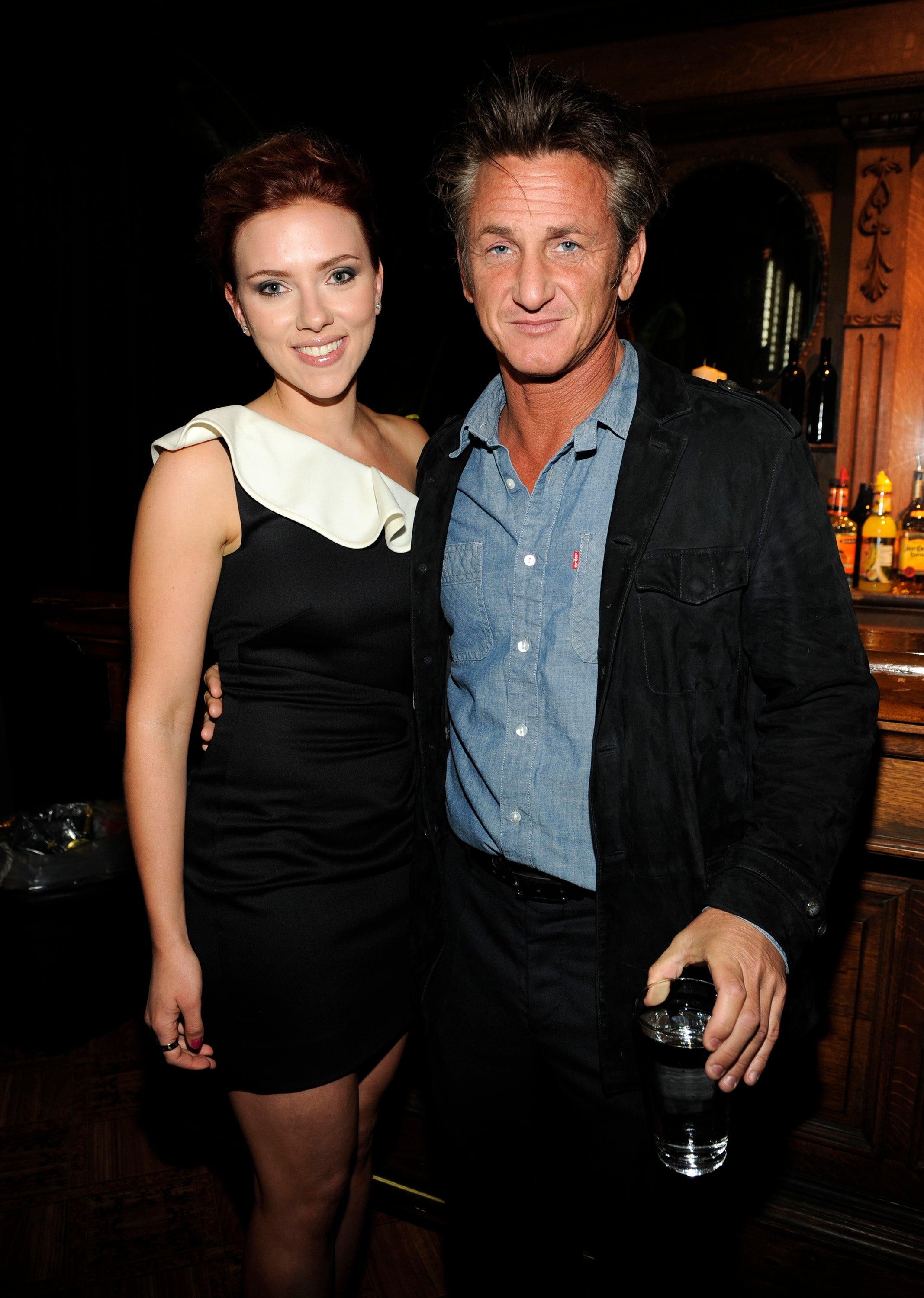 Two people pose together; one wears a stylish dress with an asymmetrical neckline, the other in a casual jacket and shirt, both smiling at an event