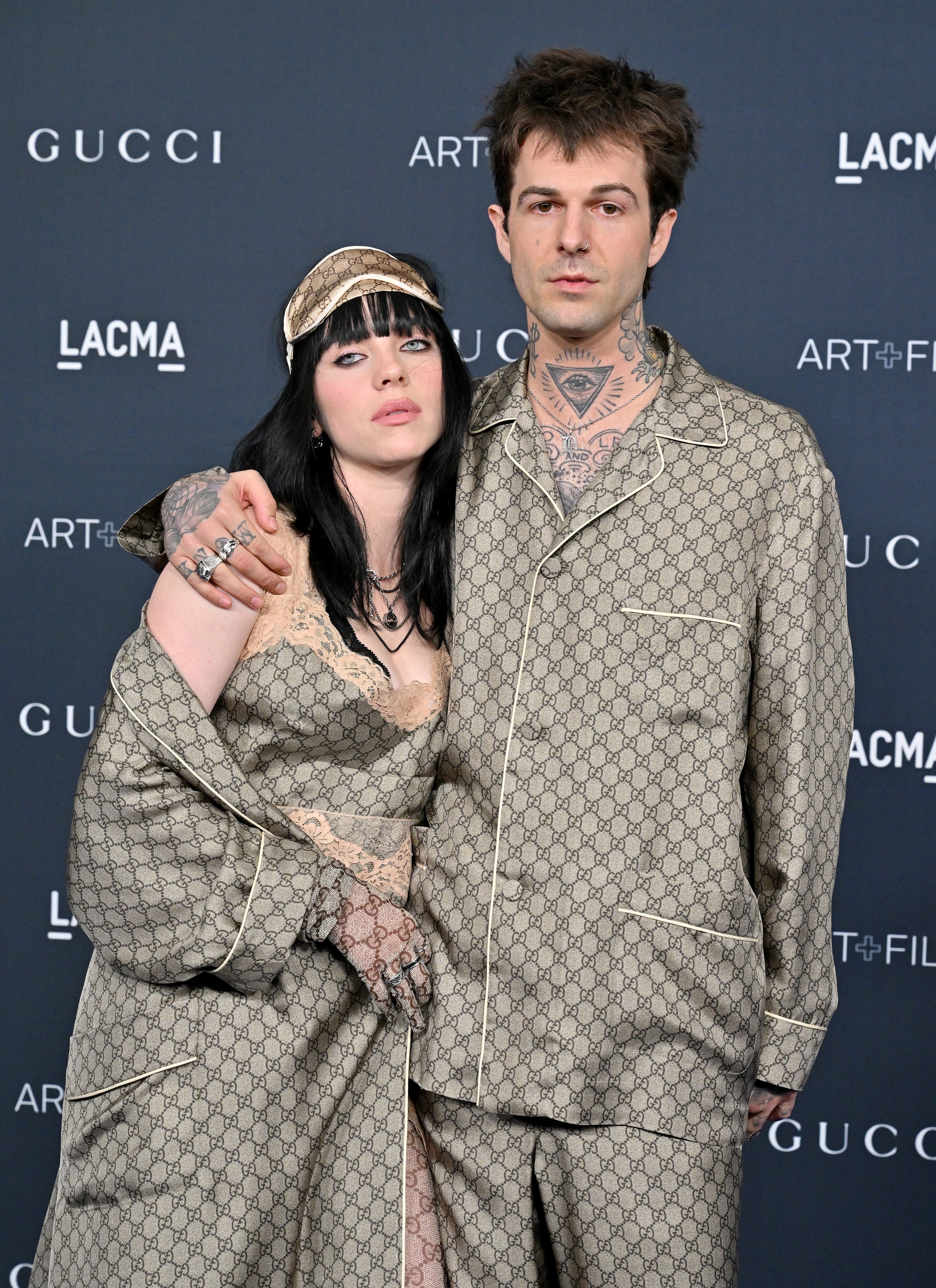 Two individuals in matching patterned outfits pose together at a Gucci event. Both wear pajama-style clothing with accessories