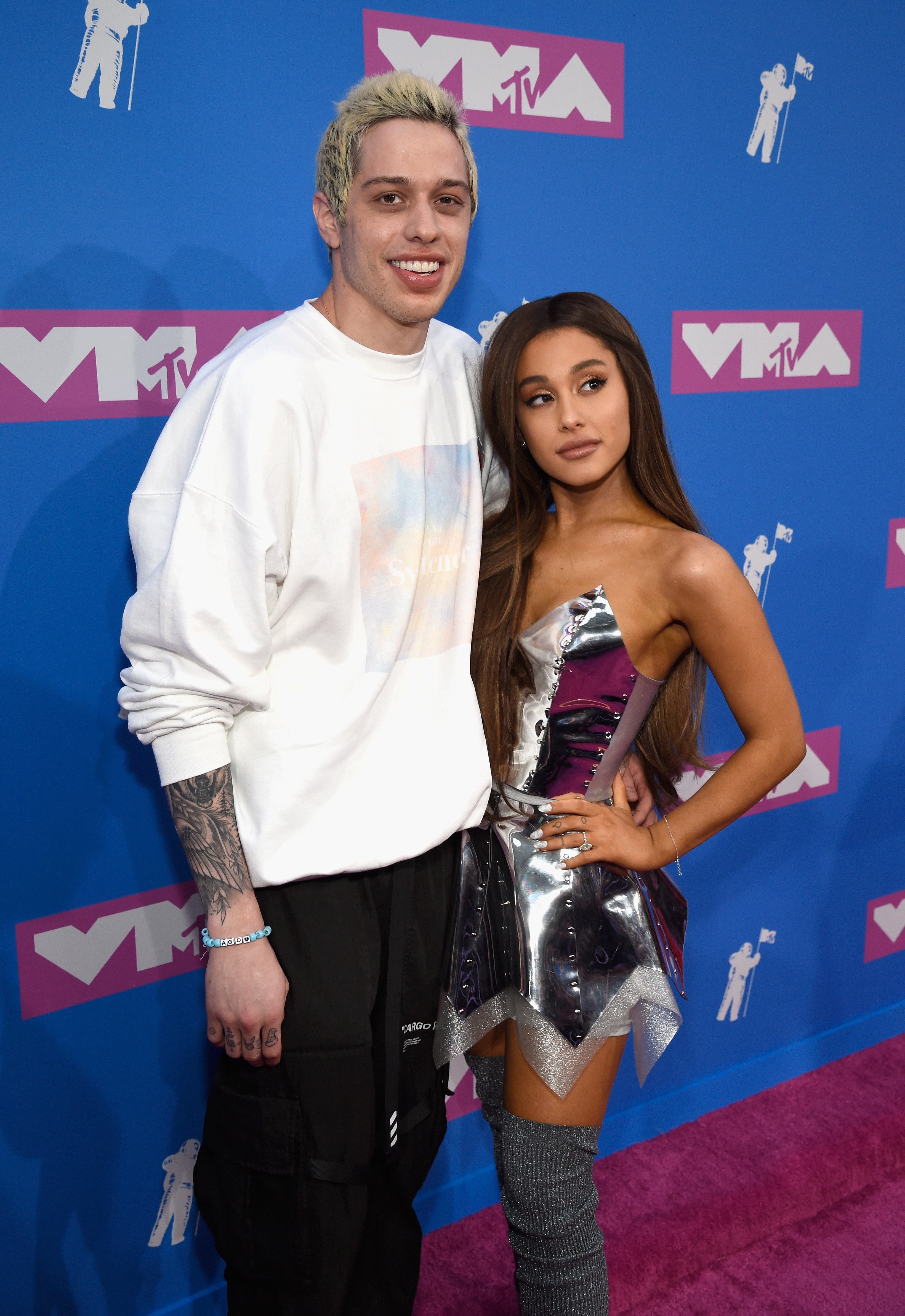 Two people posed on a red carpet; one in a casual outfit and the other in a silver metallic dress with thigh-high boots at an MTV event