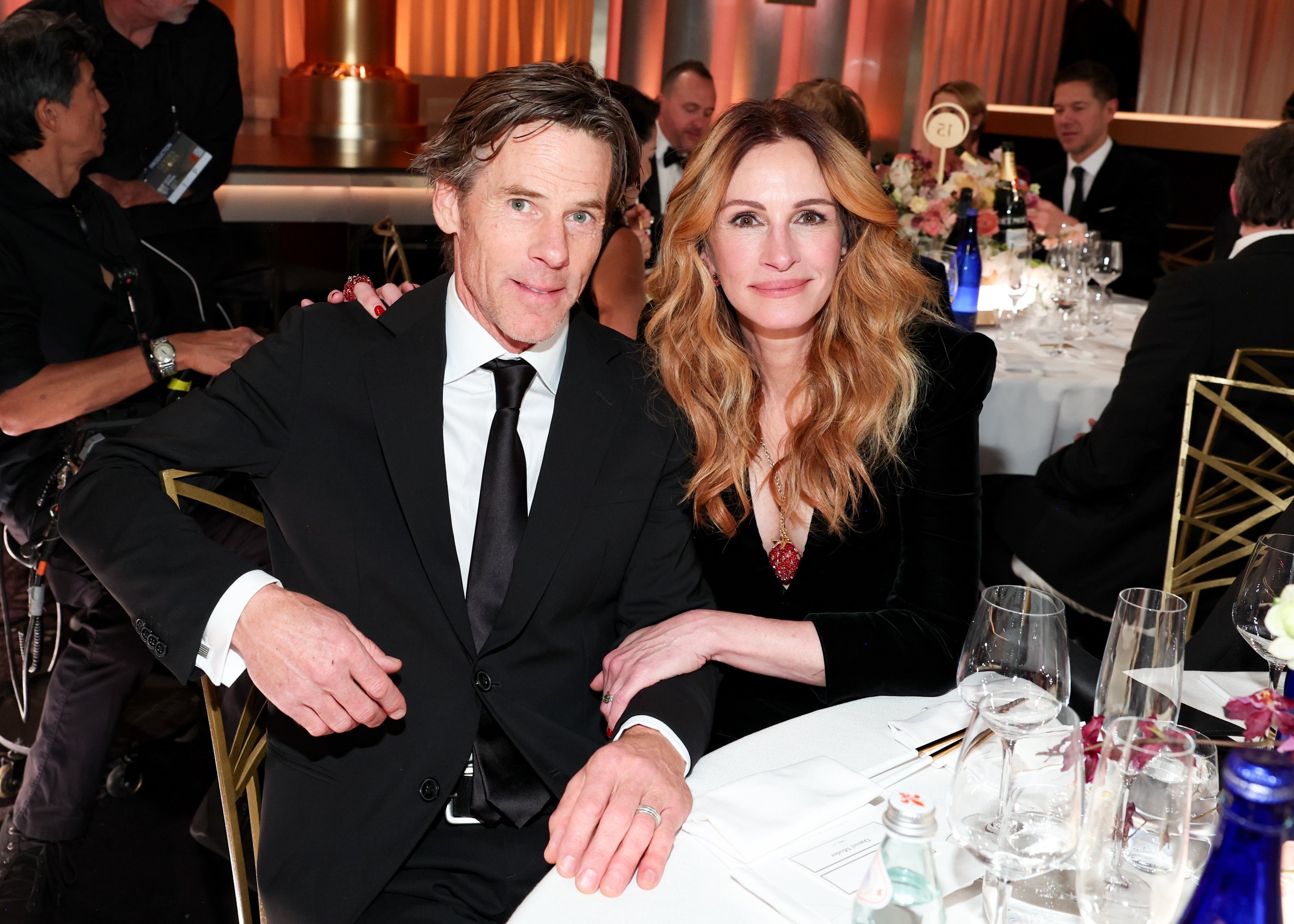 Two people seated at an event table, both in formal attire. The woman wears a black blazer with wavy hair, and they are posing for the camera