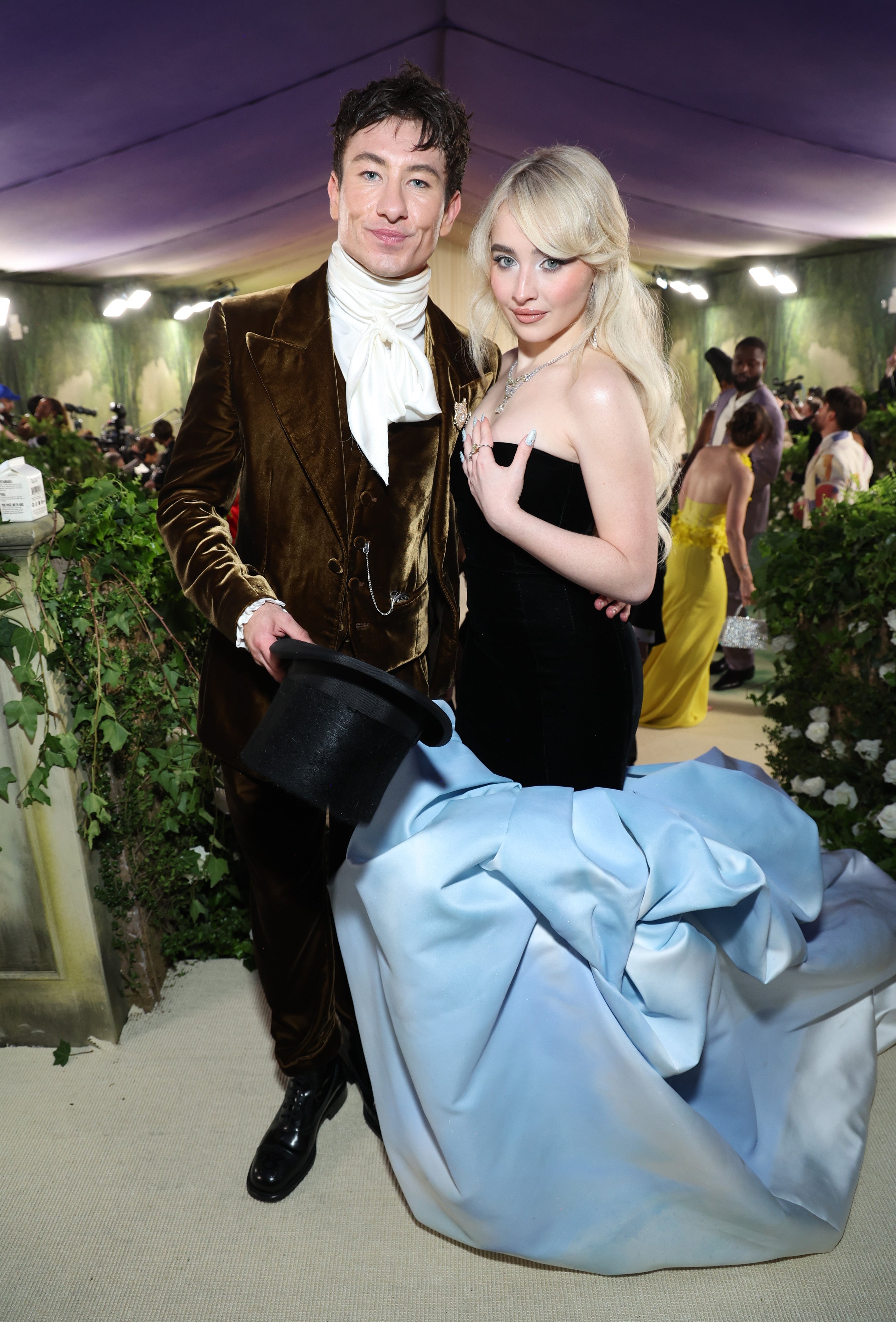 Two individuals pose at a formal event. One wears a velvet suit with a cravat, the other a strapless gown with a long train