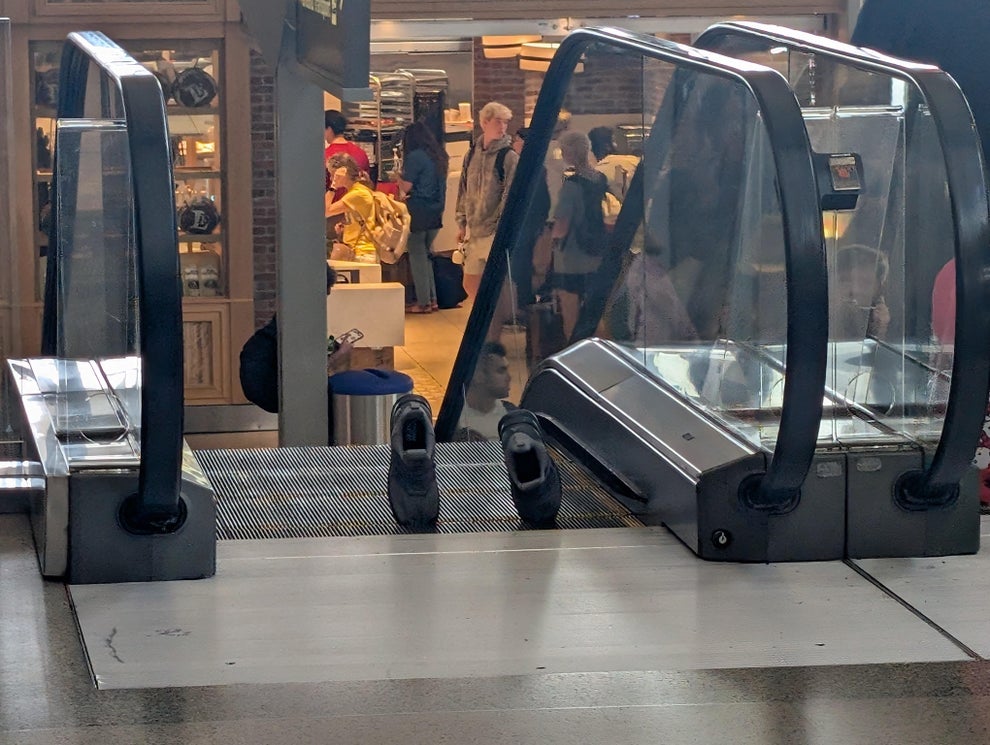 A pair of shoes stands upright at the base of an inactive escalator in a busy shopping area, with people and stores in the background