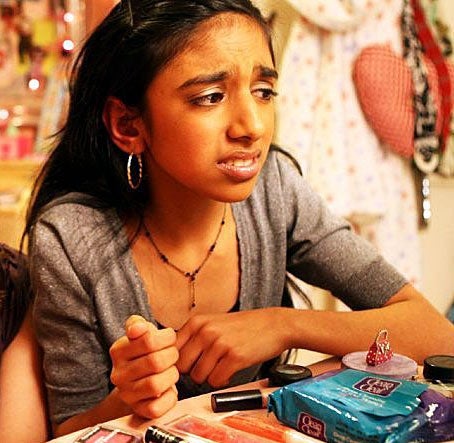 A young person sits at a vanity table with makeup products, appearing pensive with a concerned expression