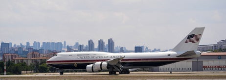 アメリカ合衆国の政府専用機が滑走路を走行。背景に都市の高層ビル群が見える。前景には数人の人々がいる。
