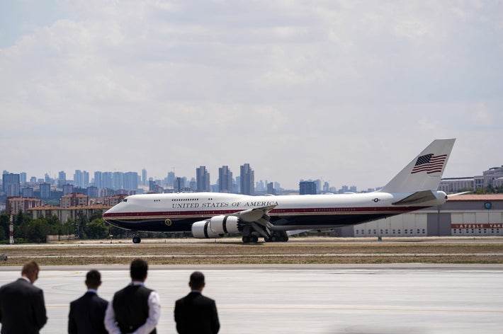アメリカ合衆国の政府専用機が滑走路を走行。背景に都市の高層ビル群が見える。前景には数人の人々がいる。