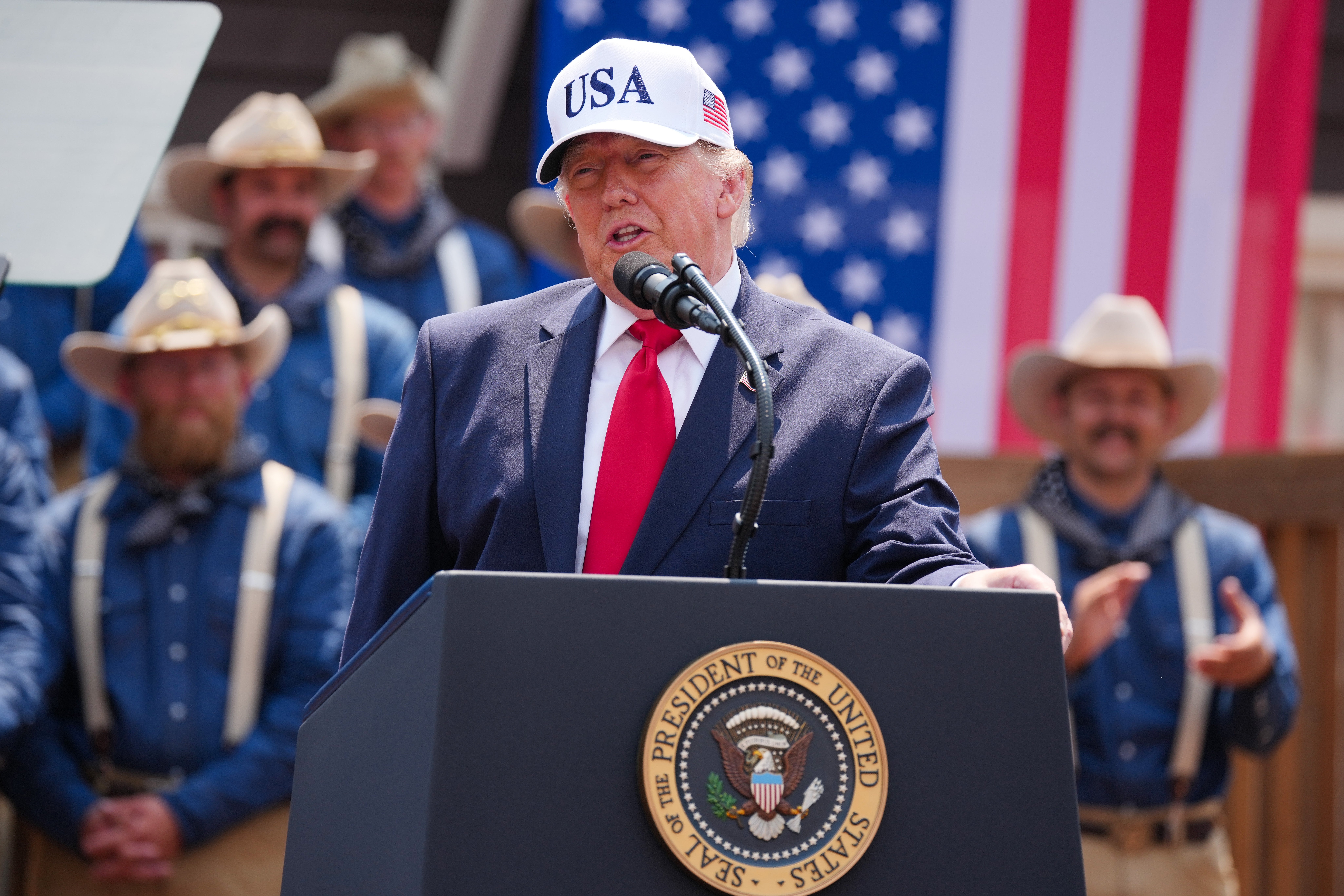 En person i et "USA" cap snakker på et podium med presidentseglet, omgitt av folk i vestlig antrekk og et amerikansk flagg i bakgrunnen