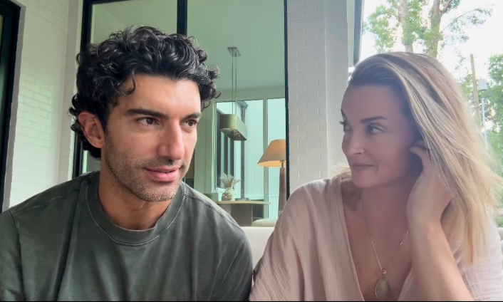 Two people sit closely together, casually dressed, in a modern indoor setting, looking at each other and appearing relaxed and engaged