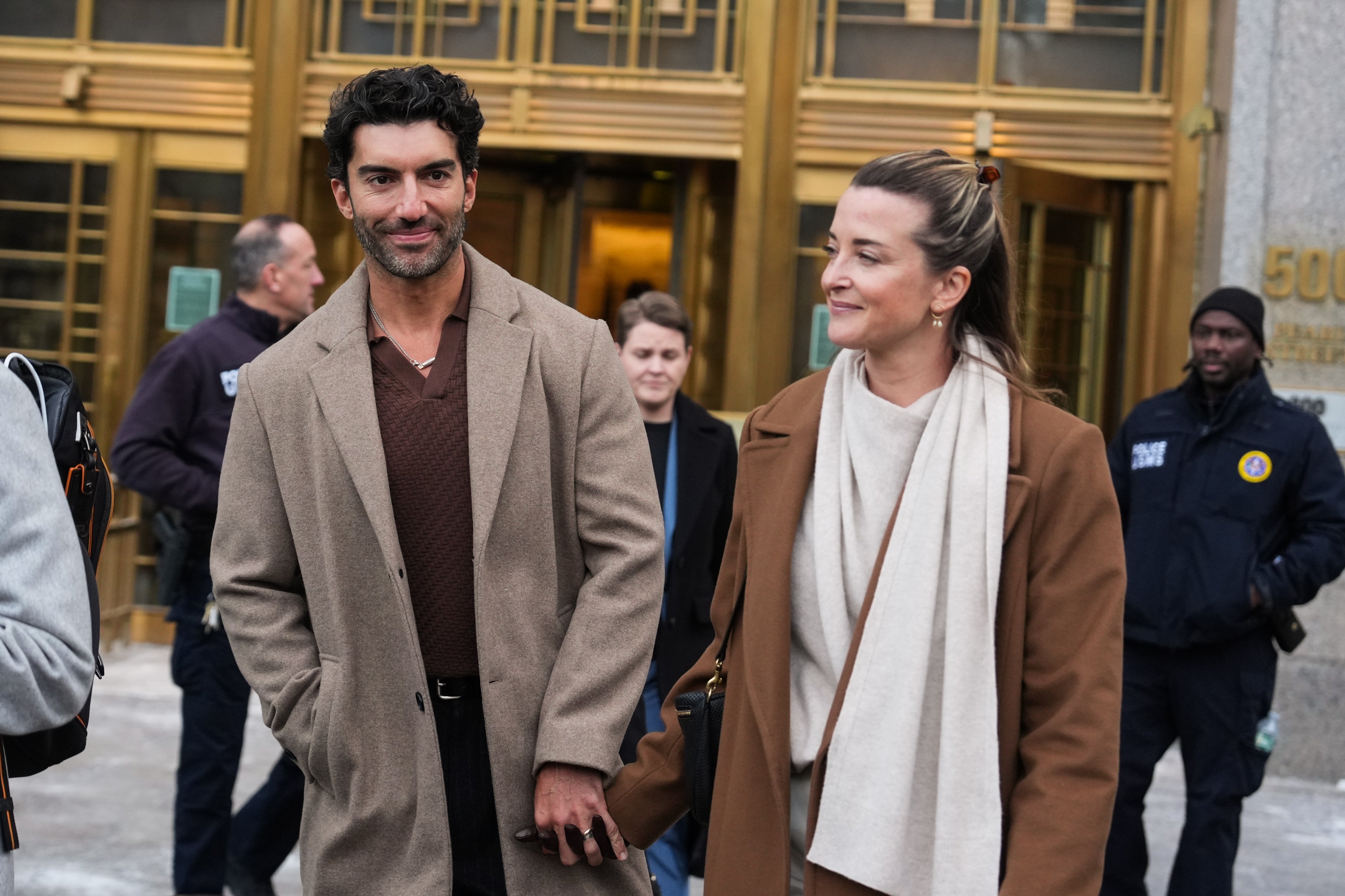 A couple is walking hand-in-hand outside a building, both wearing stylish coats. Security personnel are visible in the background