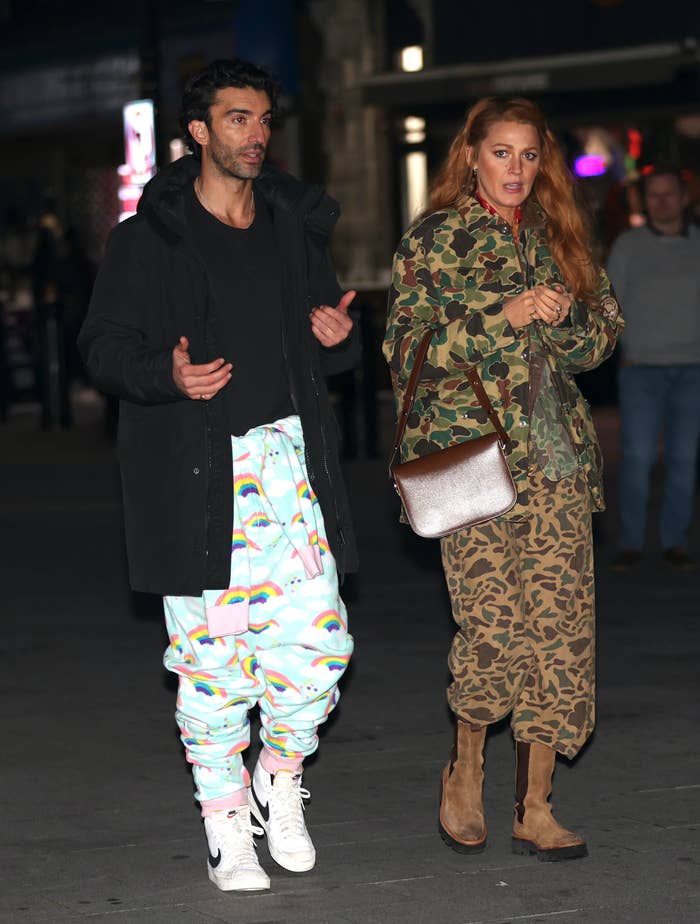 Two individuals walk together; one wears vibrant patterned pants and a black coat, the other in camouflage jacket and leopard print pants with boots