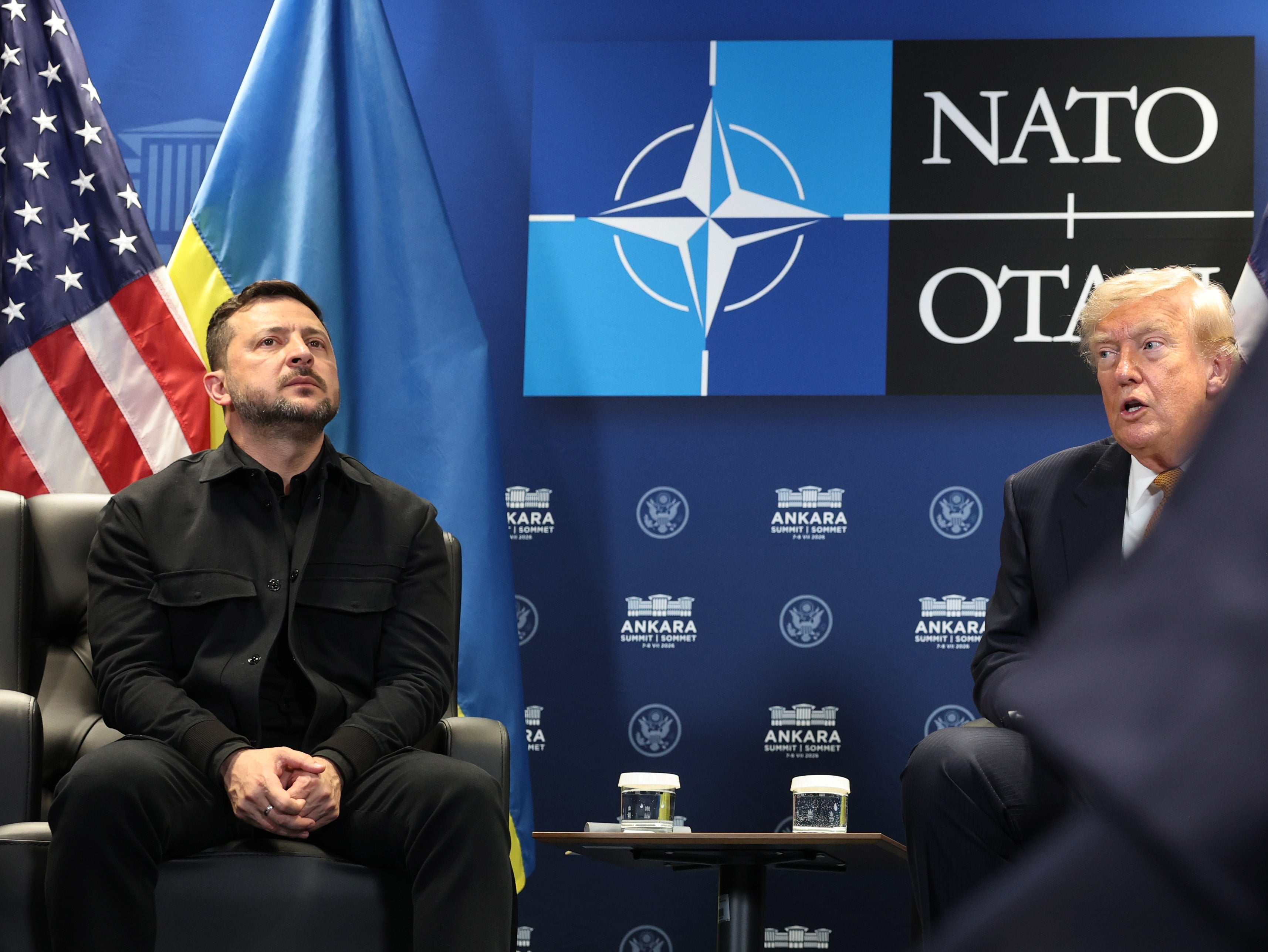 To menn sitter foran NATO-logoen, engasjert i diskusjon, med flagg i bakgrunnen