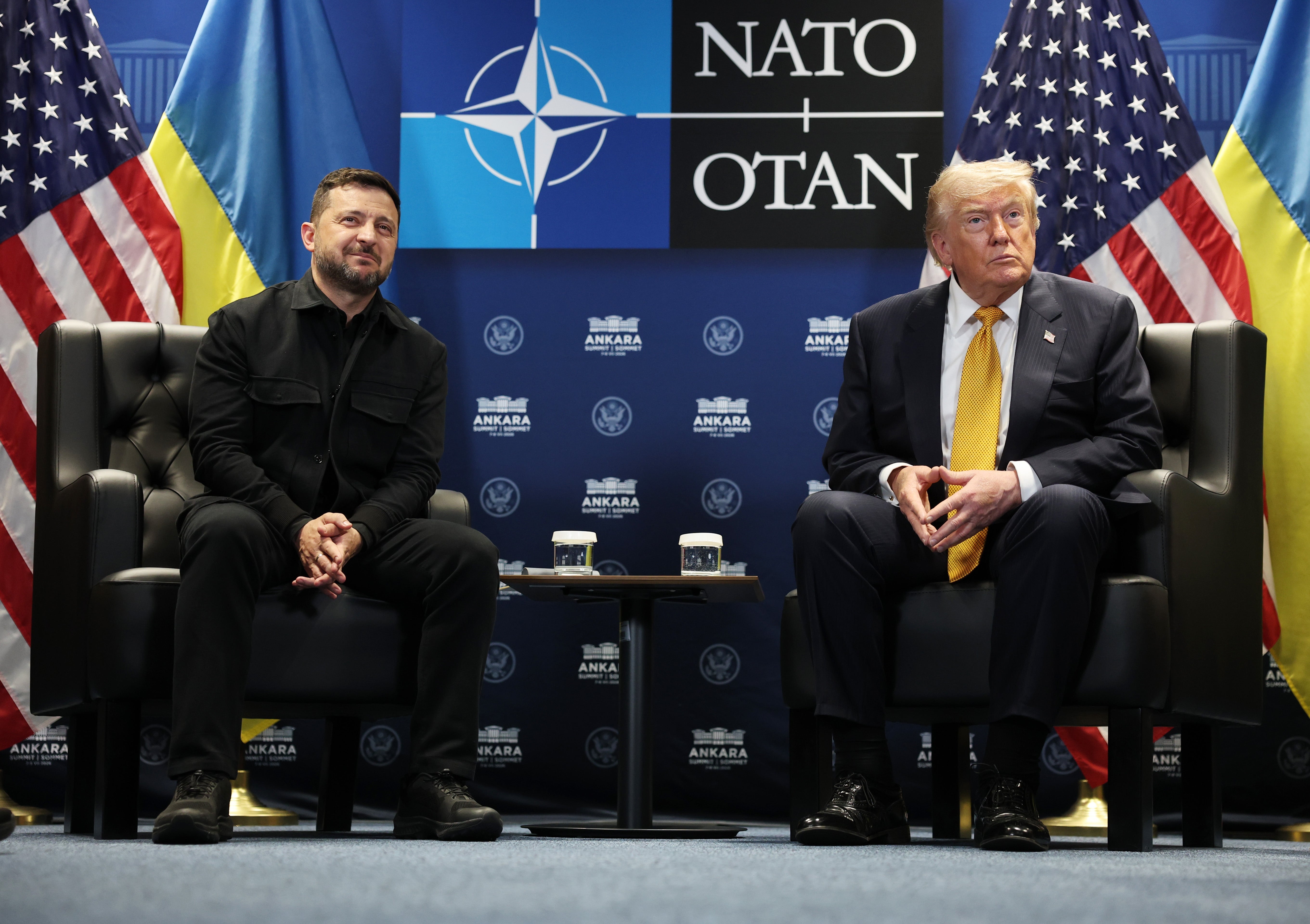 To personer sittende, en i svart dress og den andre i marinedress med gult slips, på et NATO-arrangement med flagg i bakgrunnen