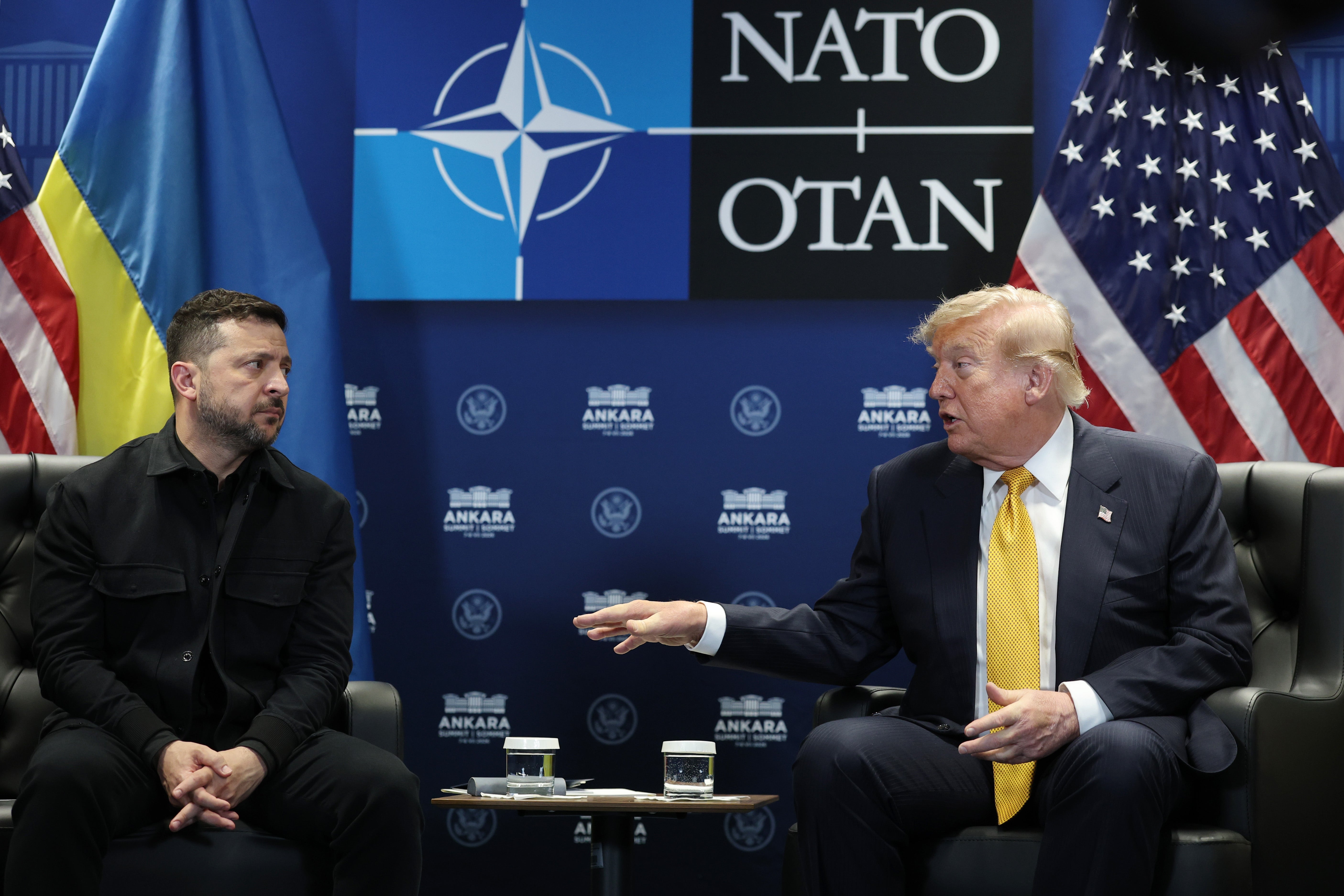 To menn i dress sitter og snakker på et NATO-arrangement, med nasjonalflagg og en NATO-logo i bakgrunnen