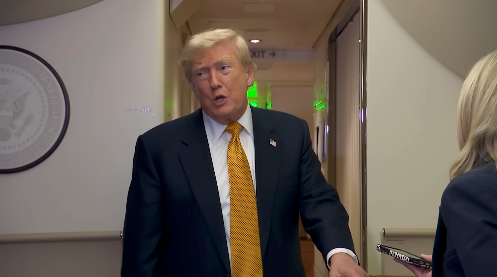 Mann i dress med oransje slips snakker til en person som holder en notatbok, inne i en gang med seglet til USAs president