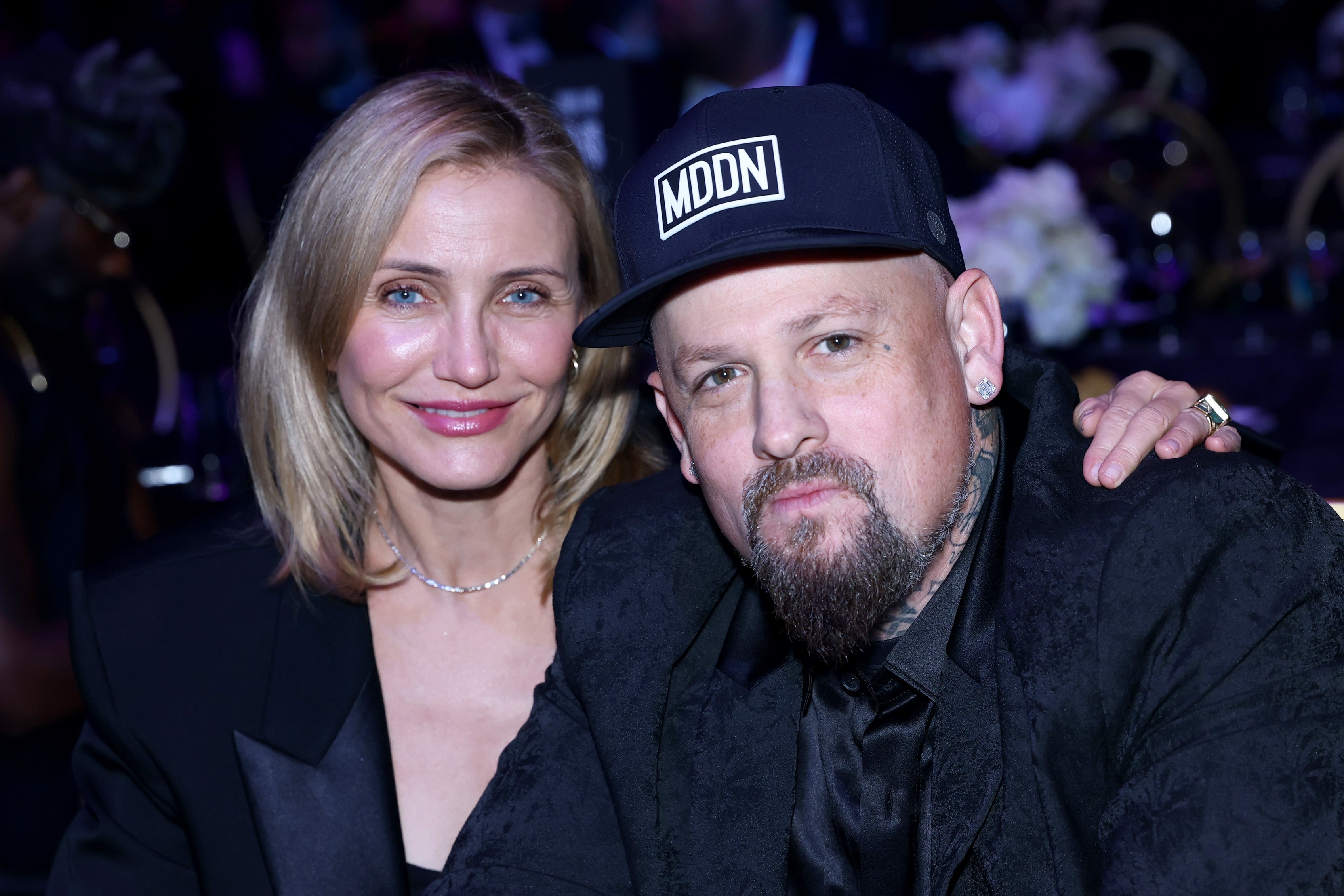 Two individuals at an event, one with shoulder-length hair in a black outfit, the other with a cap and jacket. They are posing together, smiling