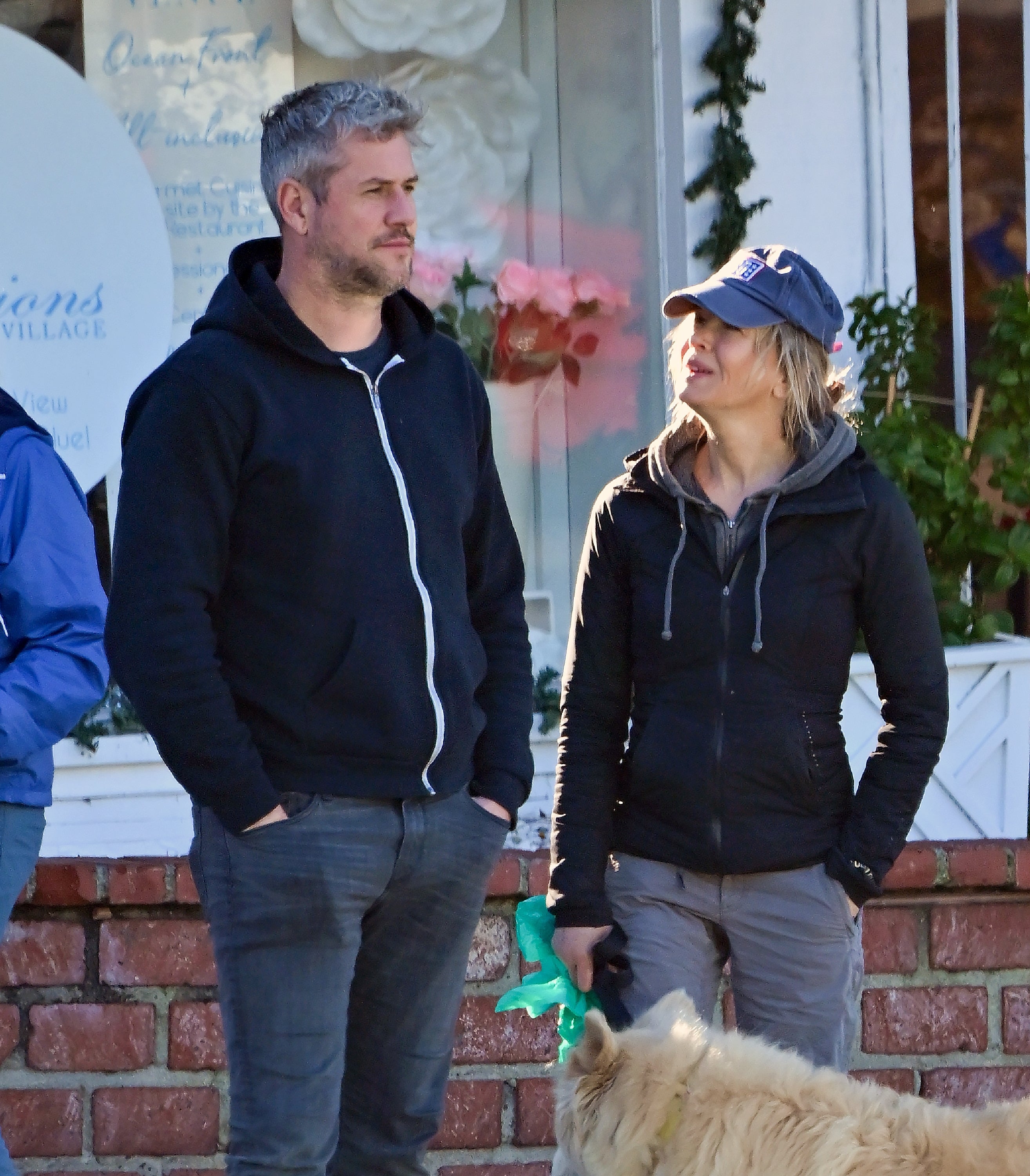 Two people casually dressed, standing outside with a dog. One wears a hoodie and jeans, the other a jacket and cap, engaged in conversation