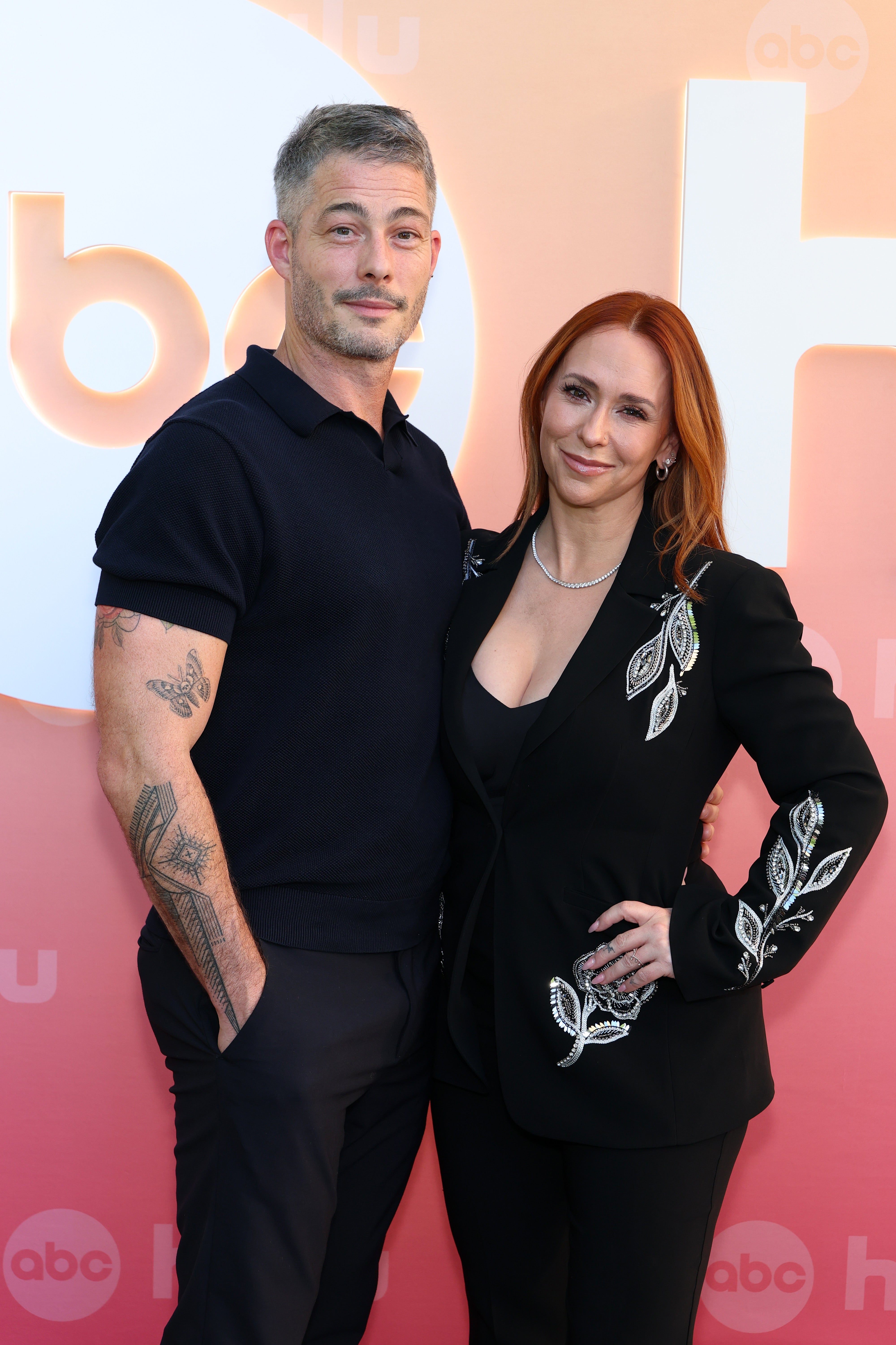 Two individuals stand on a red carpet. The person on the left is in a casual short-sleeve shirt, while the person on the right wears a decorated blazer