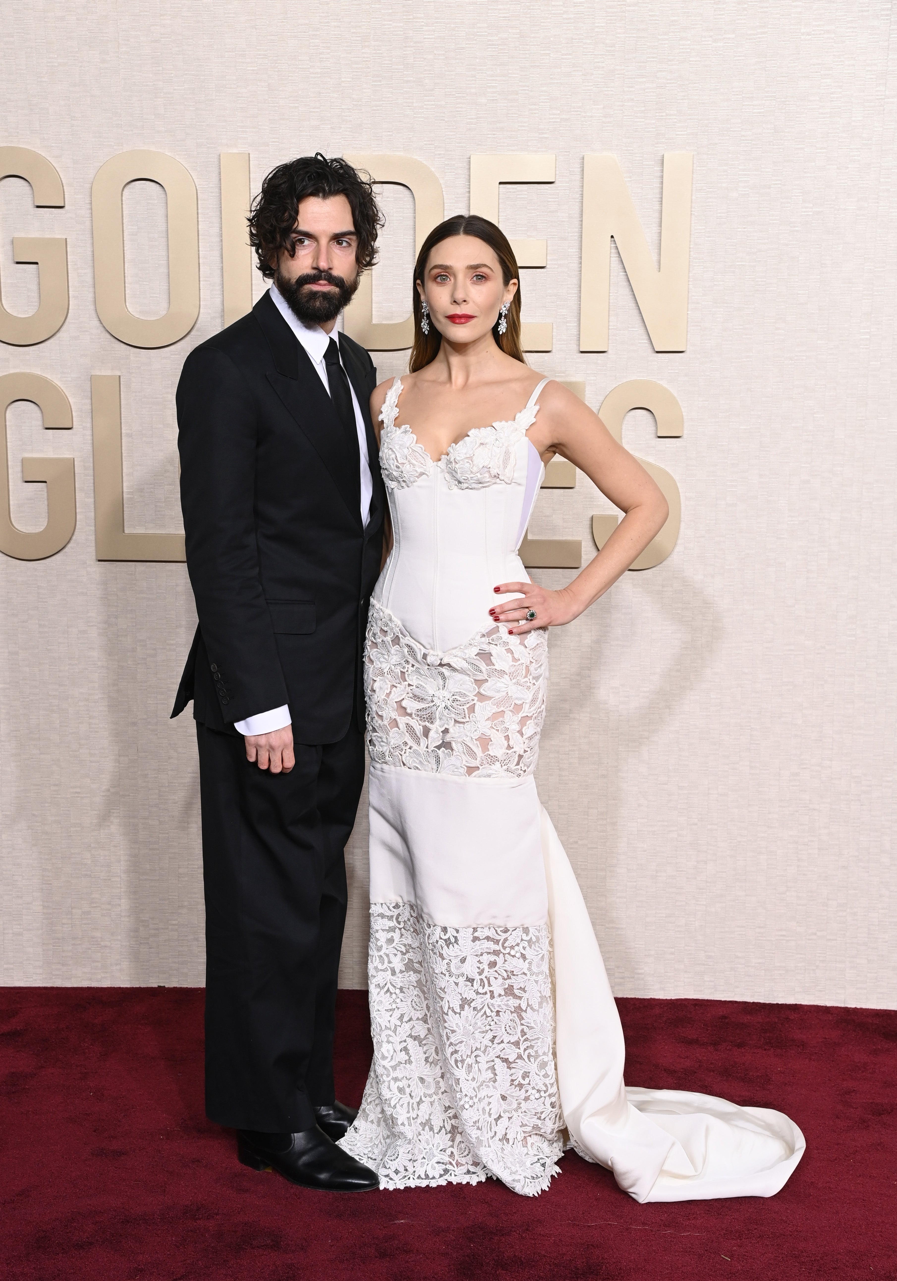 Two celebrities pose on a red carpet. The man is in a black suit, and the woman wears a form-fitting gown with intricate lace details