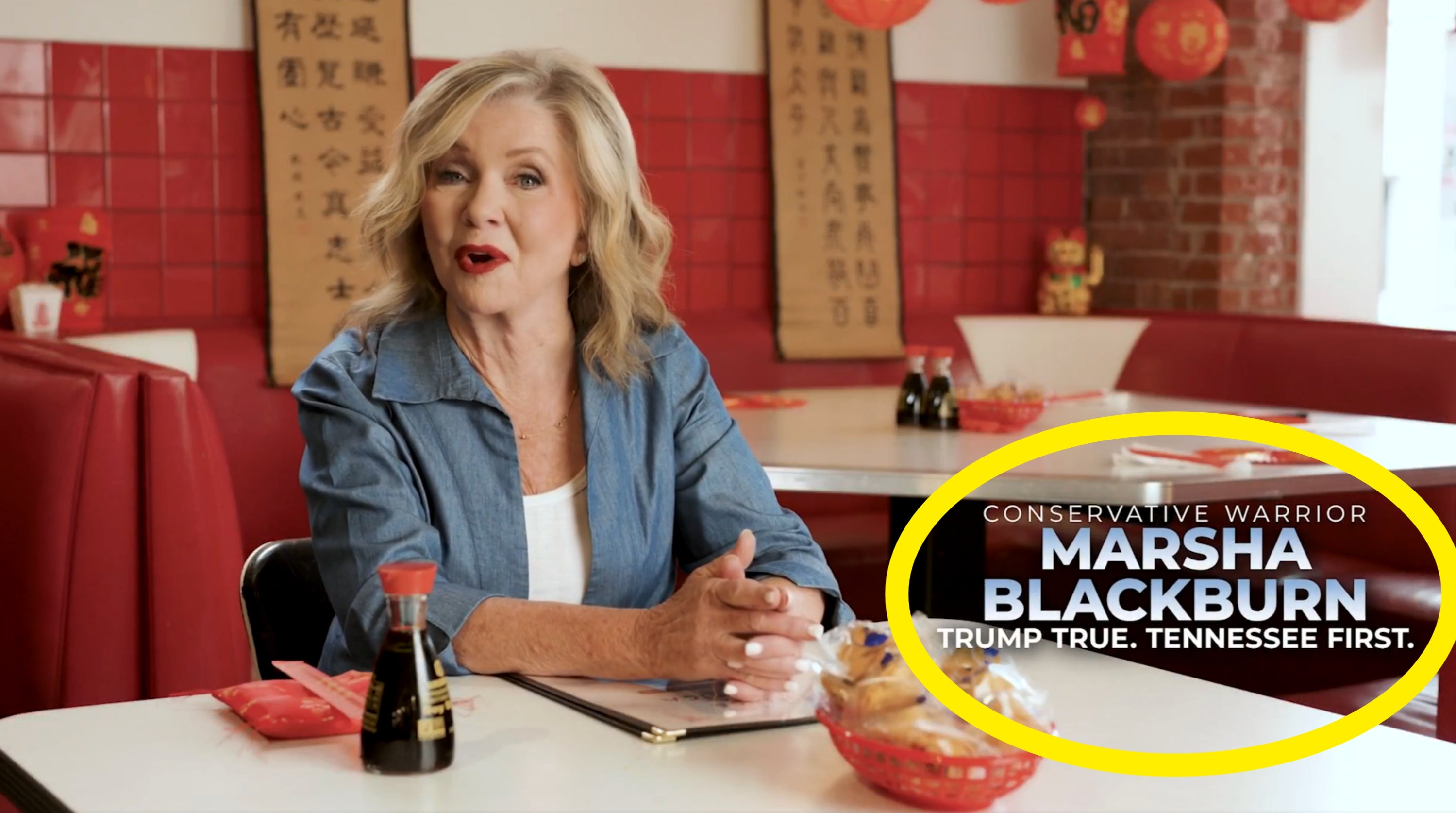En kvinne sitter ved et bord på en restaurant med røde aksenter, ved siden av en soyasausflaske og lykkekaker. Tekst: "Konservativ kriger Marsha Blackburn."