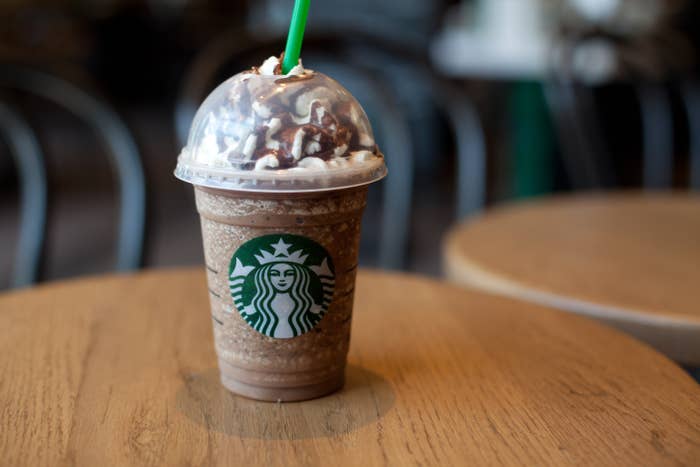 A Starbucks Frappuccino with whipped cream and chocolate drizzle on a wooden table, with empty chairs in the blurred background