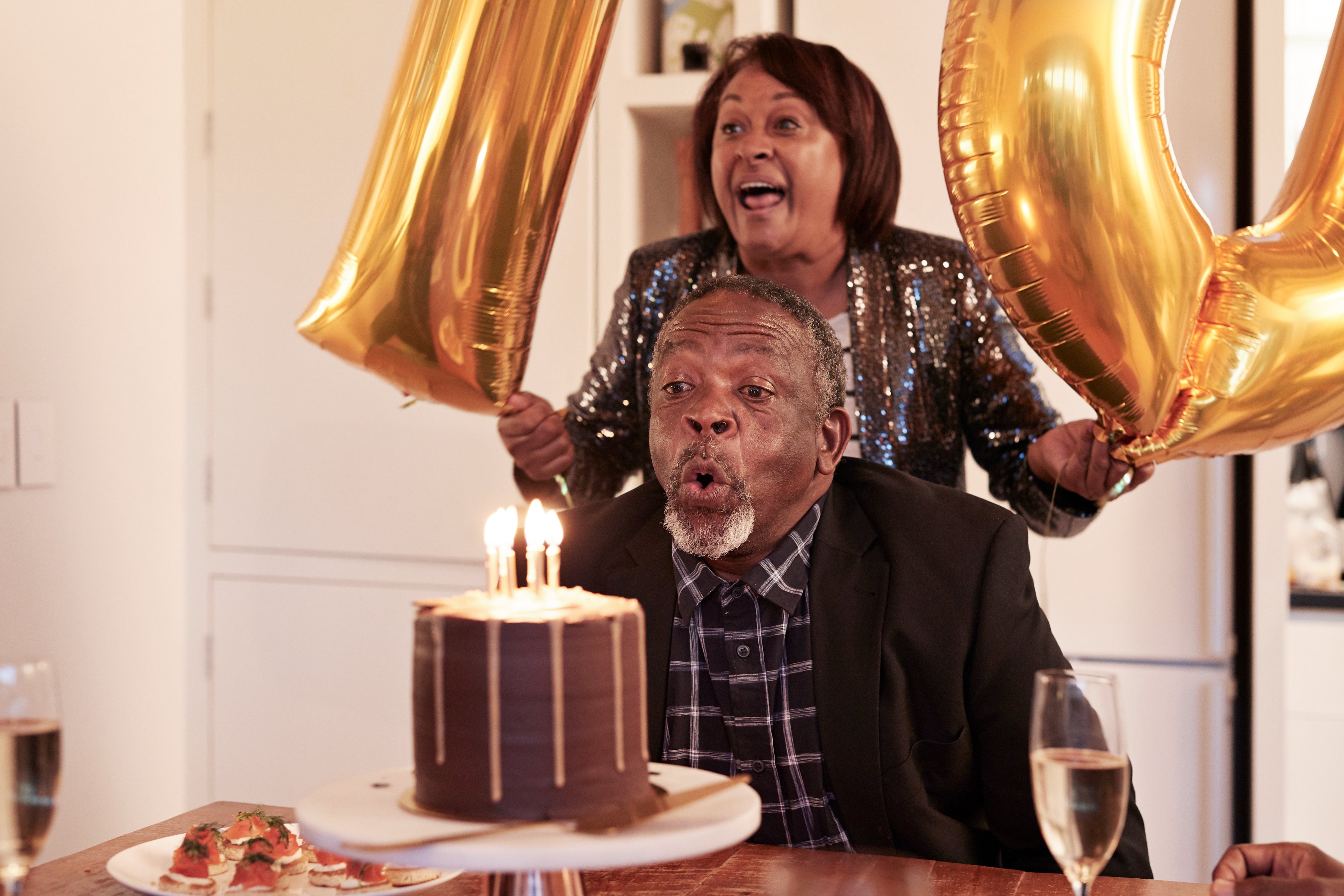 Older man blowing out candles on a cake, as a woman holds large gold balloons in the background, celebrating joyfully