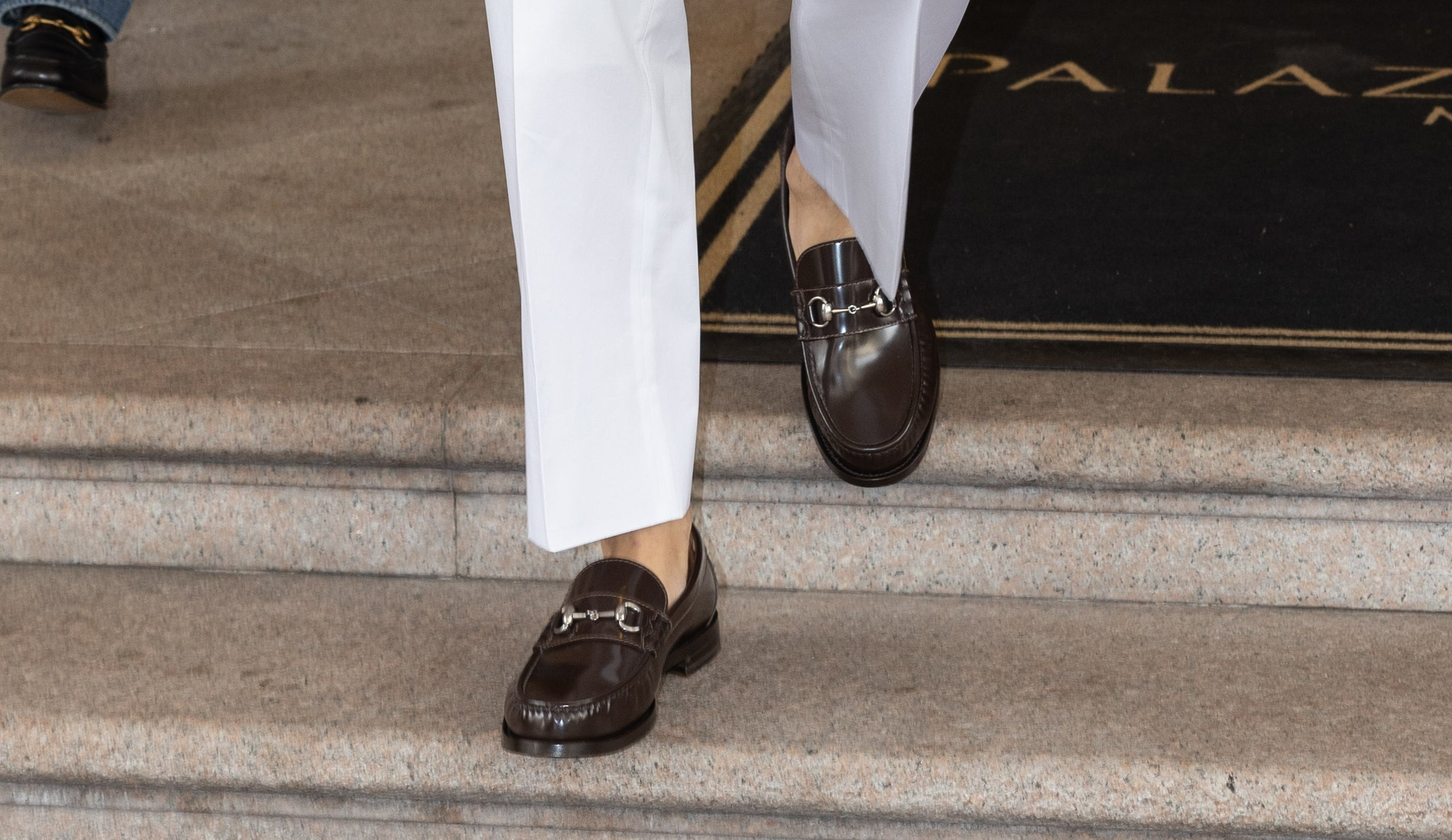 A man in a stylish white suit and dark sunglasses walks confidently down steps, followed by two others near a hotel entrance
