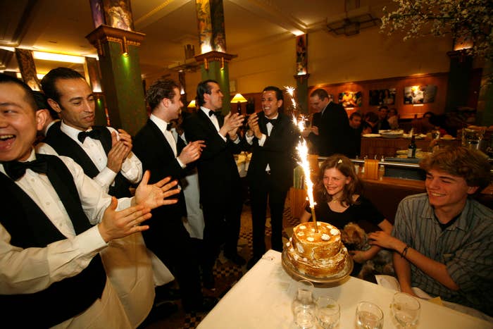 A group of people in formal attire celebrate with a birthday cake topped with sparklers in a lively restaurant setting