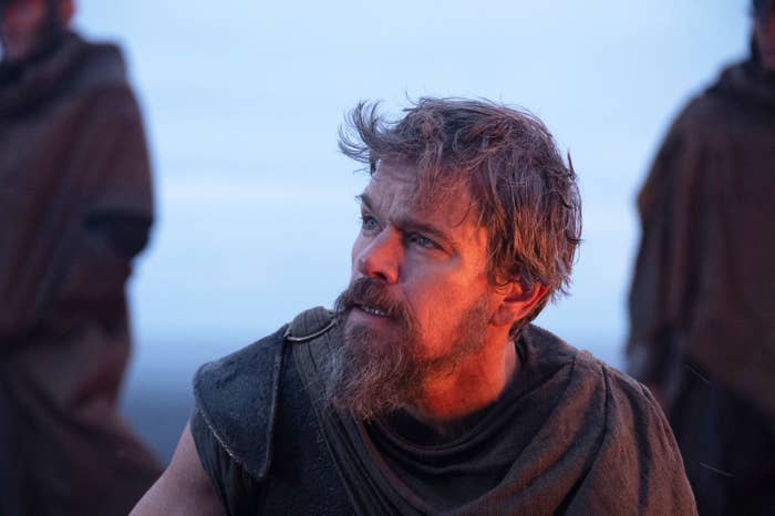 Bearded man in rugged, medieval-style clothing looks to the side, appearing to be outdoors with others in similar attire in the background