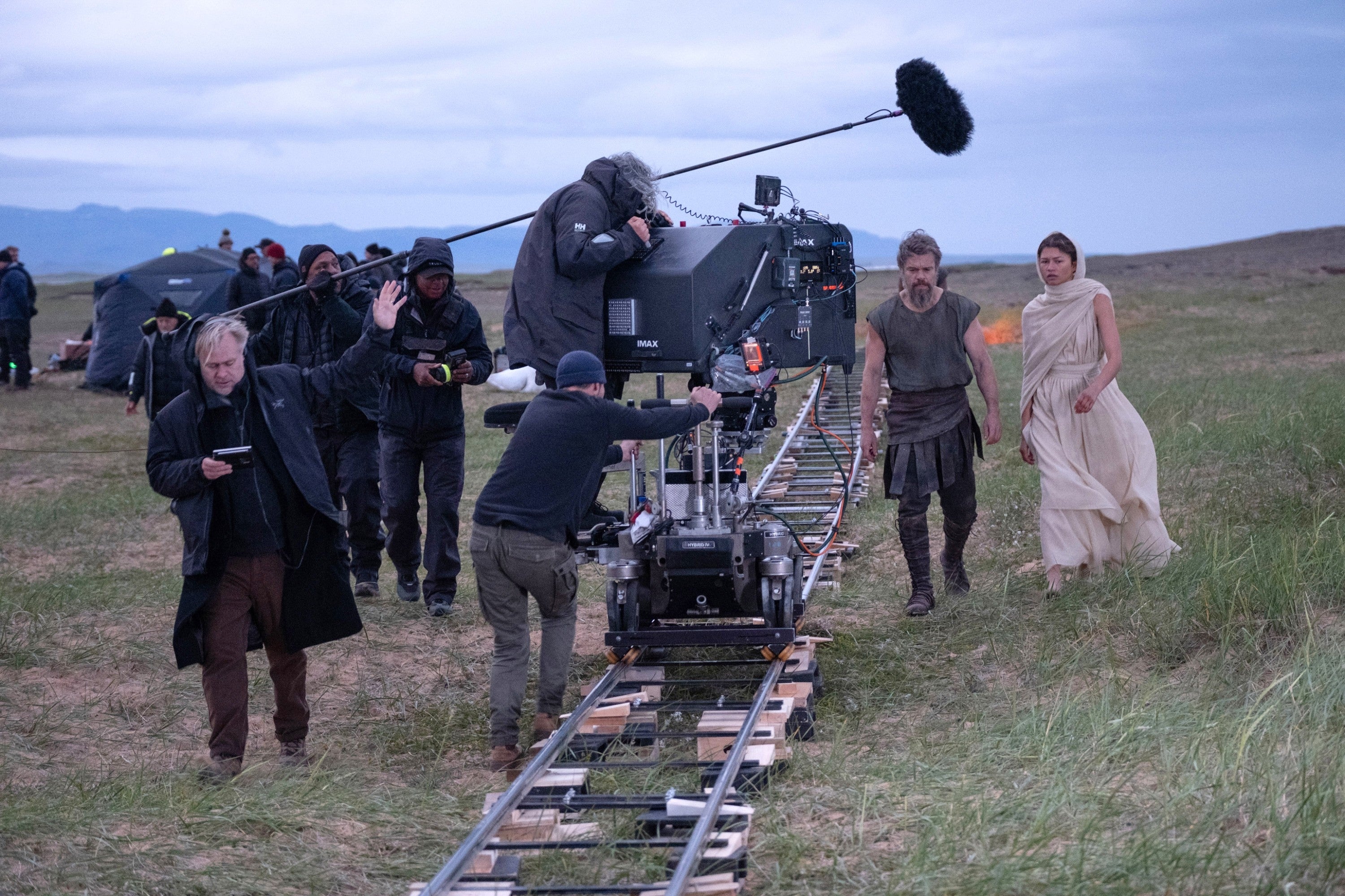 crew operates a large camera on tracks in a grassy field; an actor in rugged costume and an actress in a flowing dress observe