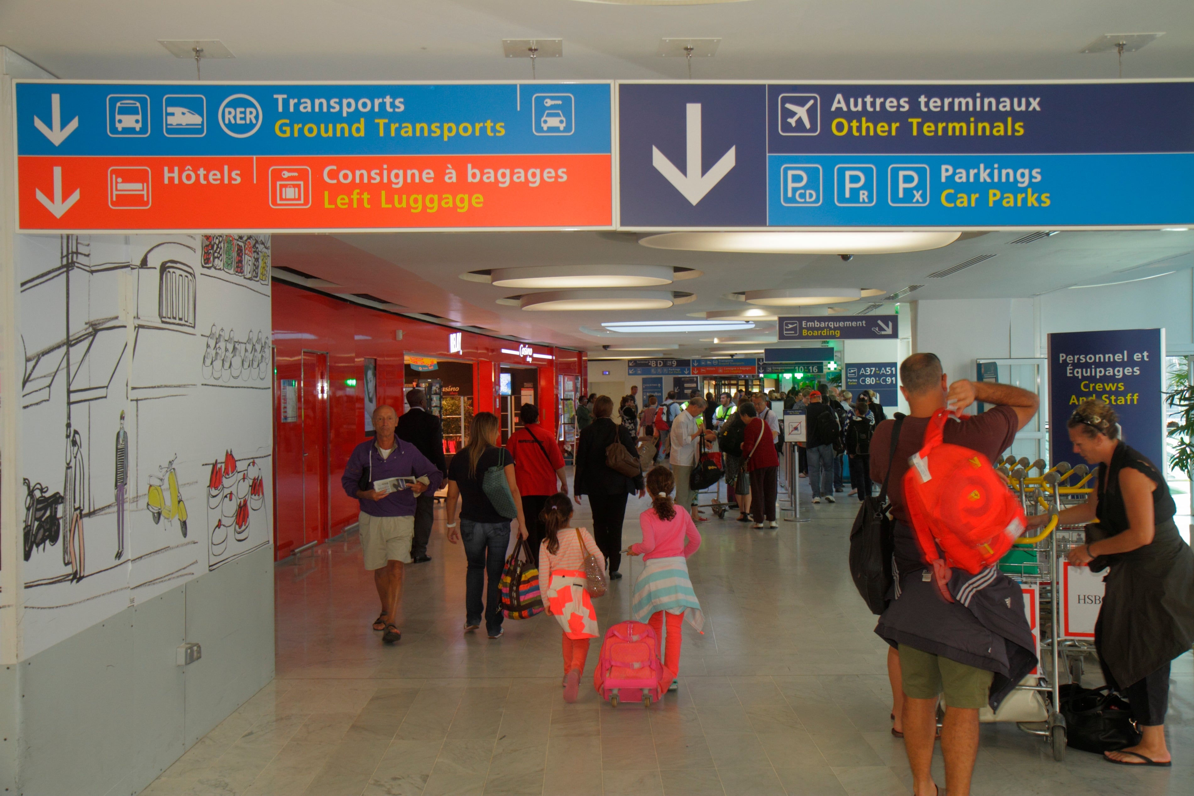 Busy airport terminal with travelers and directional signage for ground transport, hotels, left luggage, terminals, and car parks. Families and passengers navigate the area