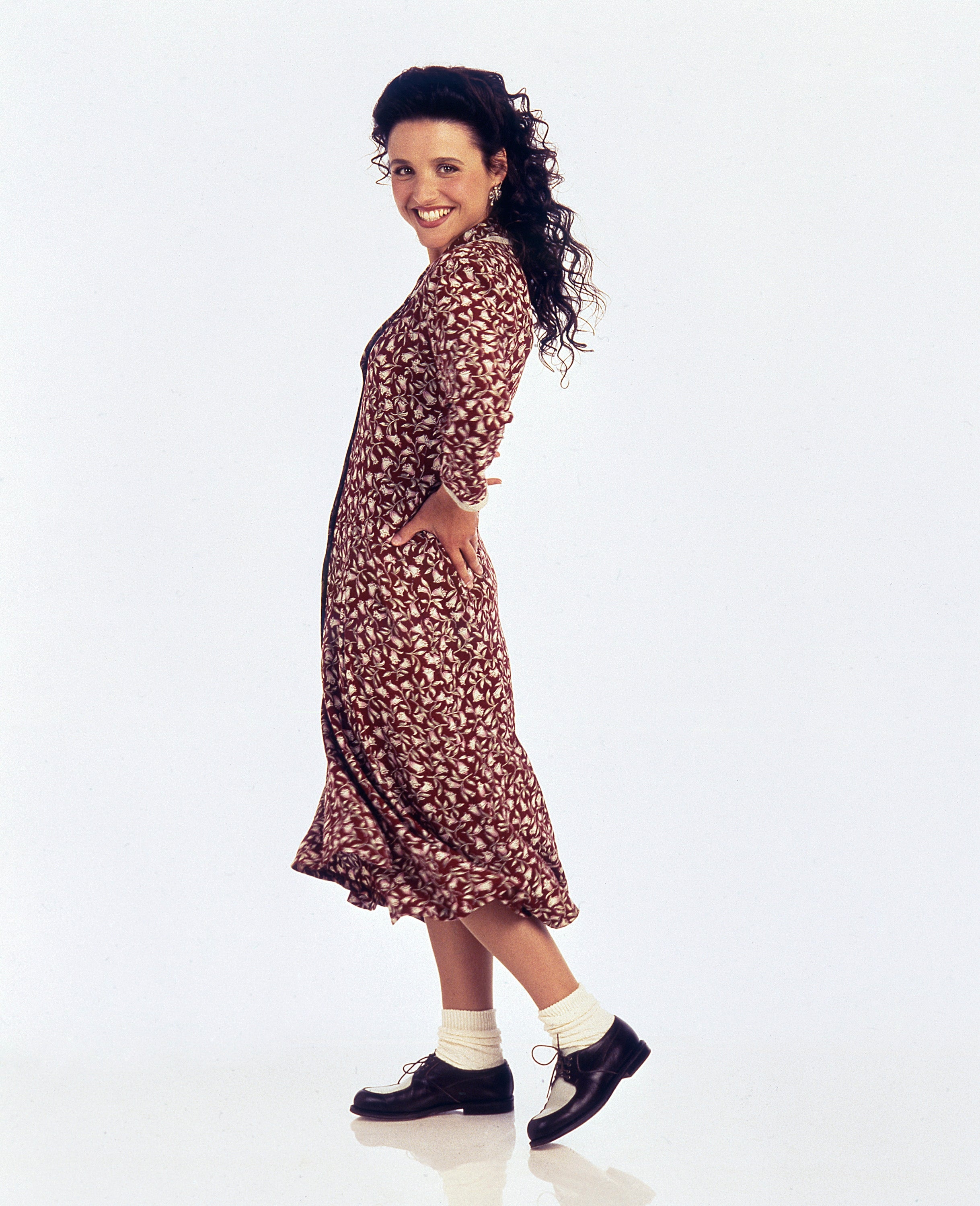 Person in a patterned dress poses playfully with hands on hips, wearing ankle socks and loafers