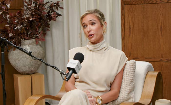 A person in an elegant, high-neck outfit sits in a cozy chair, speaking into a microphone during an interview or podcast recording