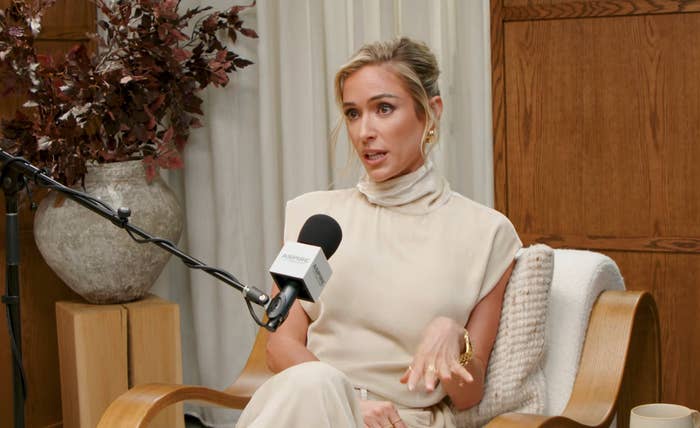 Person sitting in a chair, speaking into a microphone during a podcast interview, wearing an elegant, sleeveless high-neck outfit